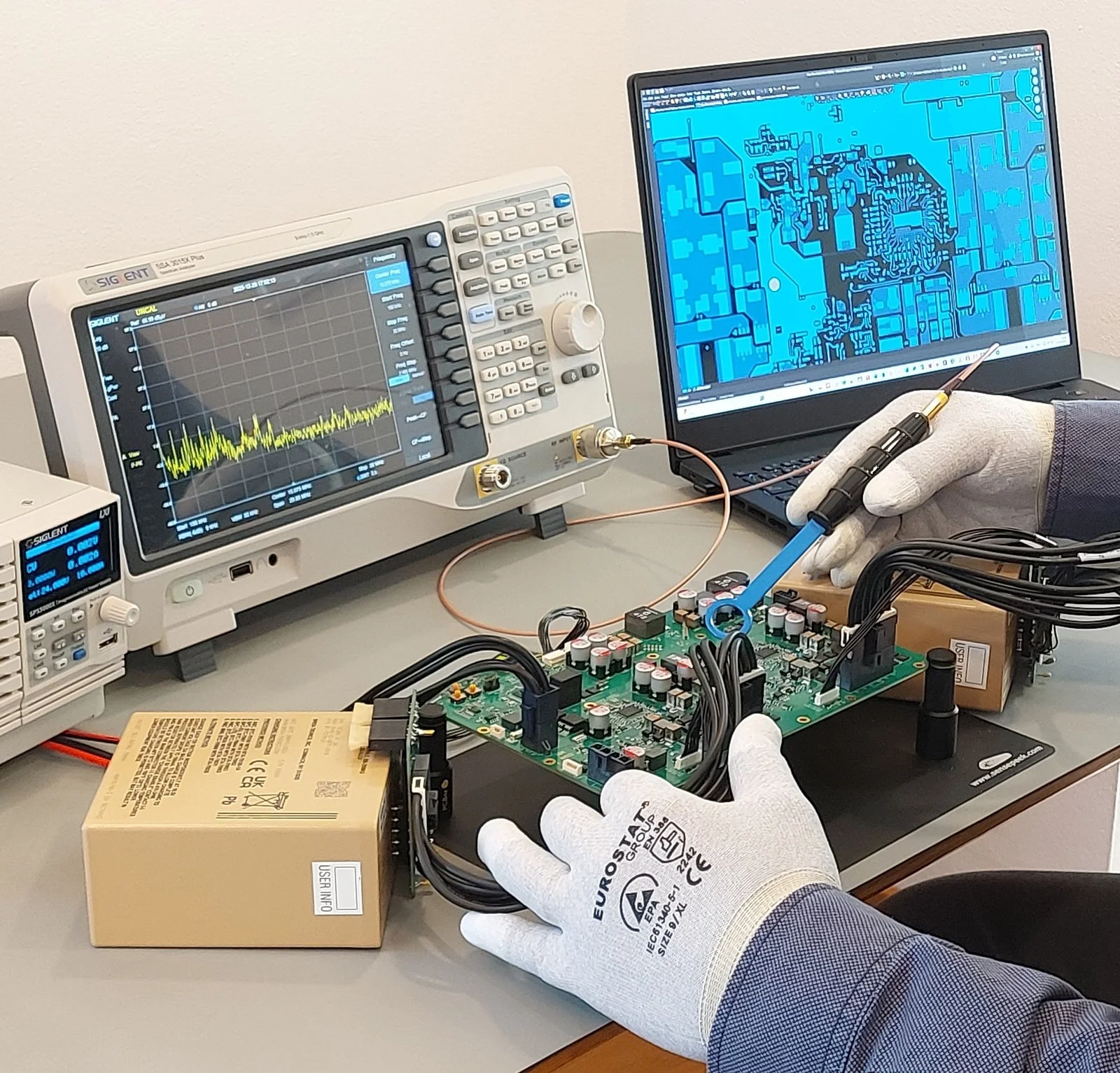 Electronic engineer measuring EMC/EMI on custom PCB board with near-field probe and spectrum analyzer