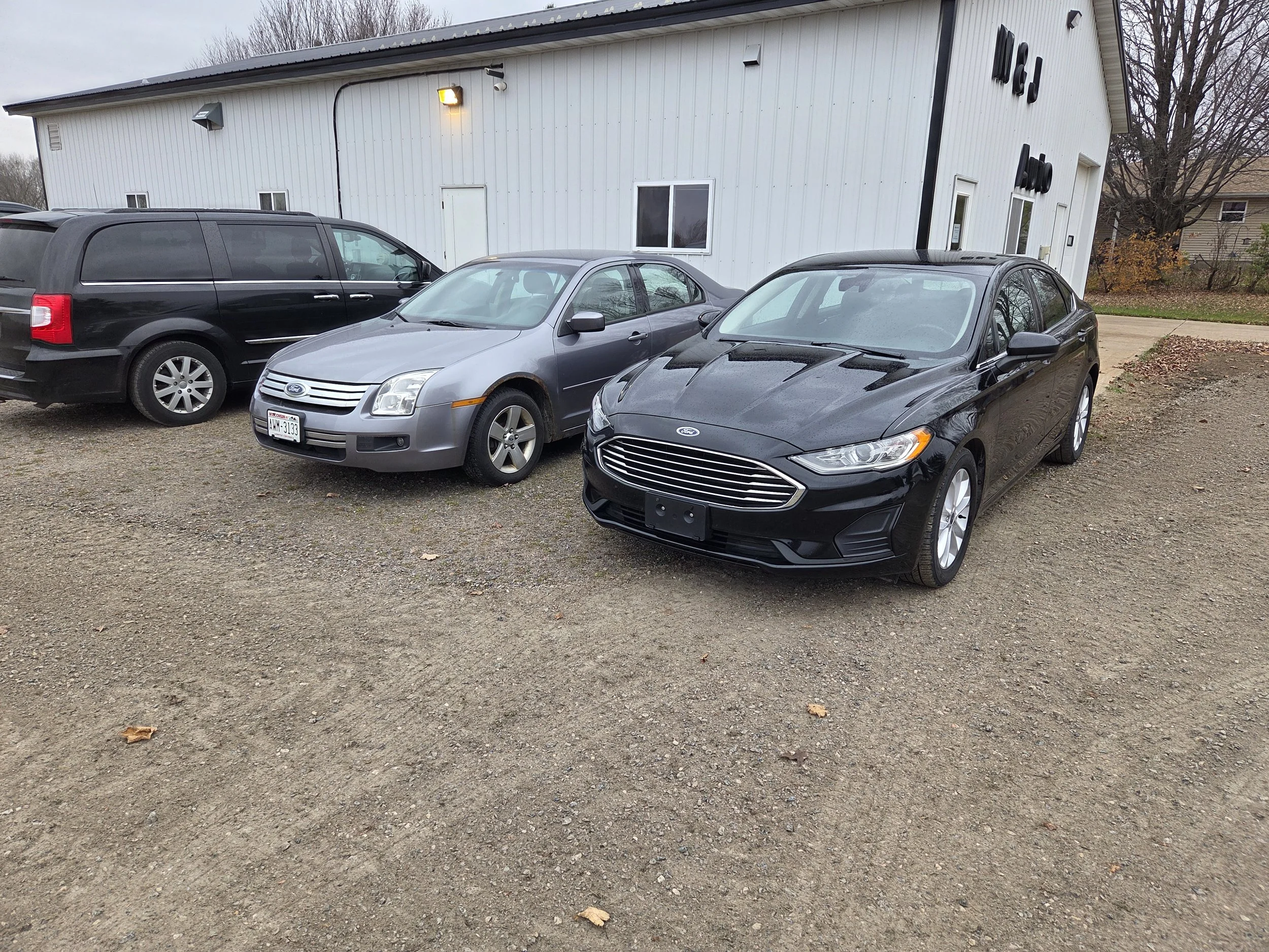 Four parked cars outside a white building on a gravel lot, including black, silver, and dark gray sedans and minivans, with trees in the background.