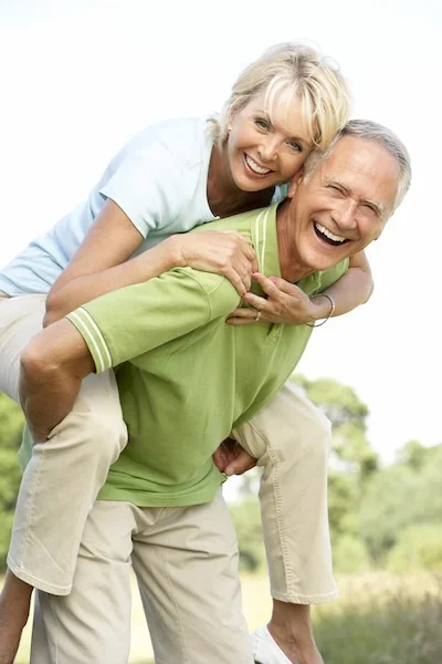 A happy elderly couple outdoors, with the woman on the man's back, all smiling and enjoying each other's company.
