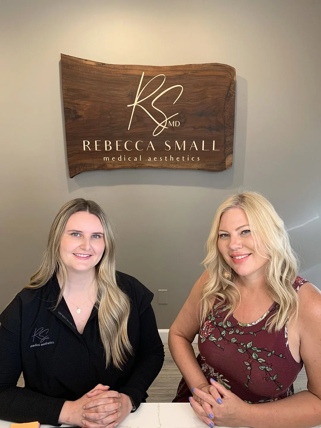 Two women sitting at a desk in front of a sign that reads 'Rebecca Small Medical Aesthetics'.