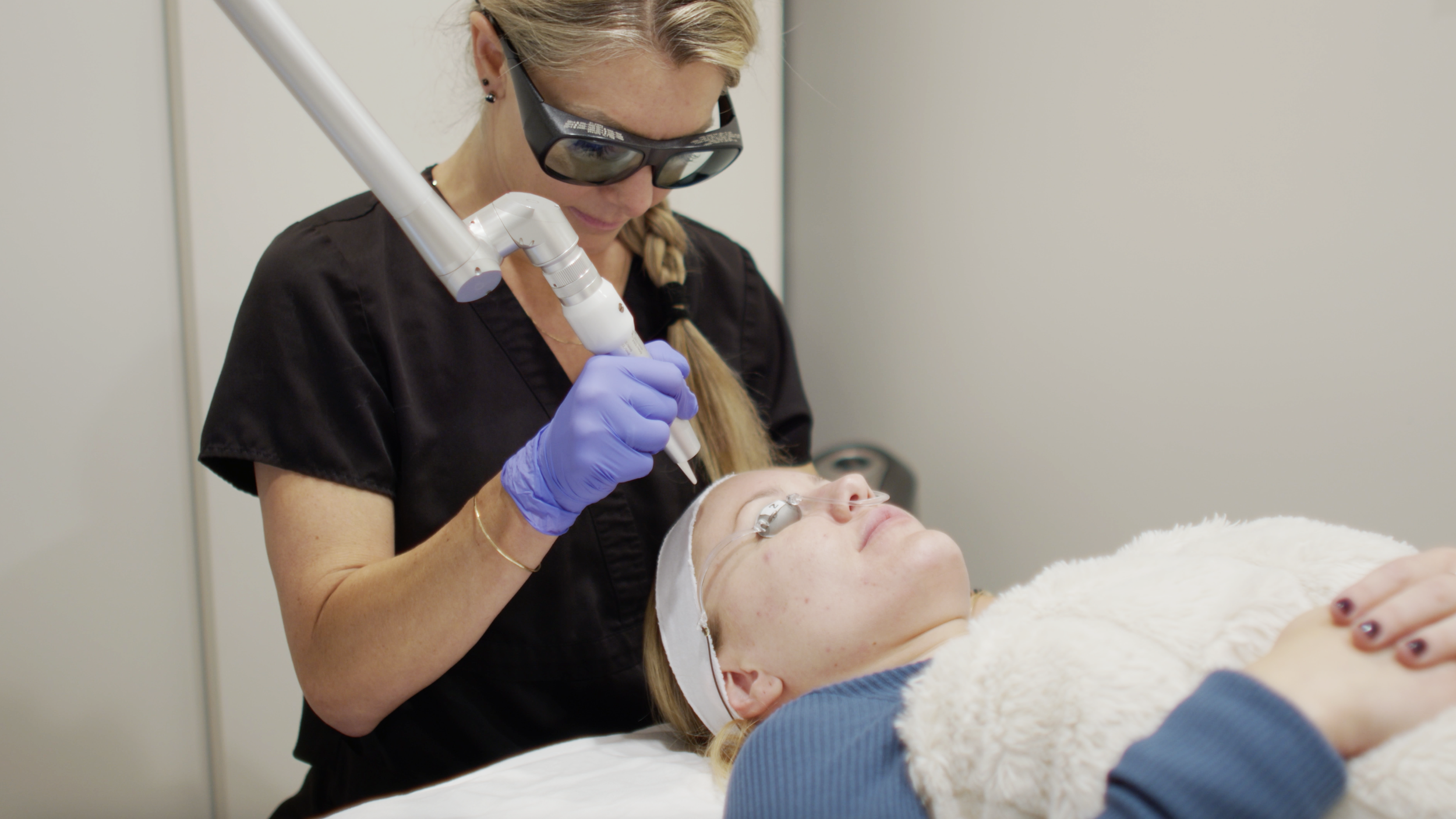 A medical professional performing a skin treatment with a laser device on a patient lying on a bed with protective eyewear.