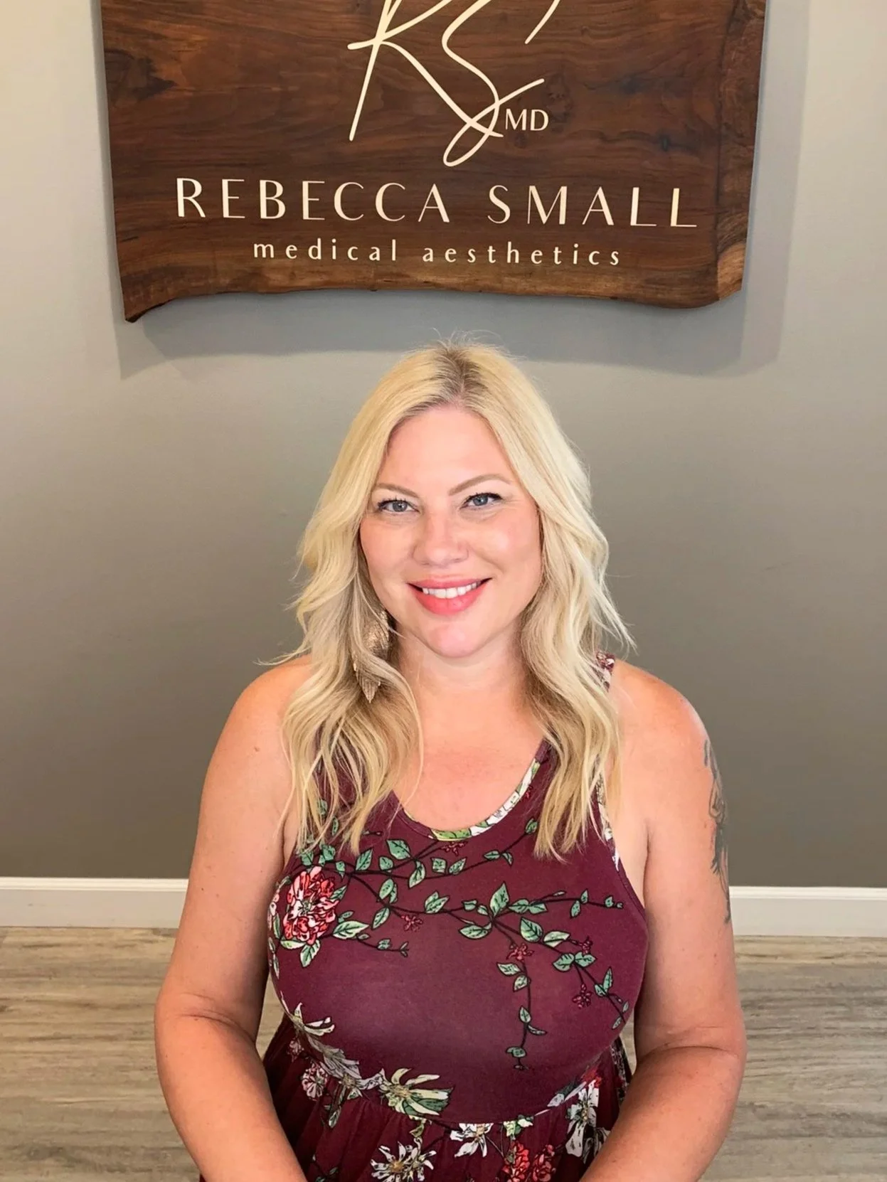 A smiling woman with blonde hair sitting in front of a sign that reads 'Rebecca Small Medical Aesthetics'. The woman is wearing a sleeveless maroon dress with a floral pattern.