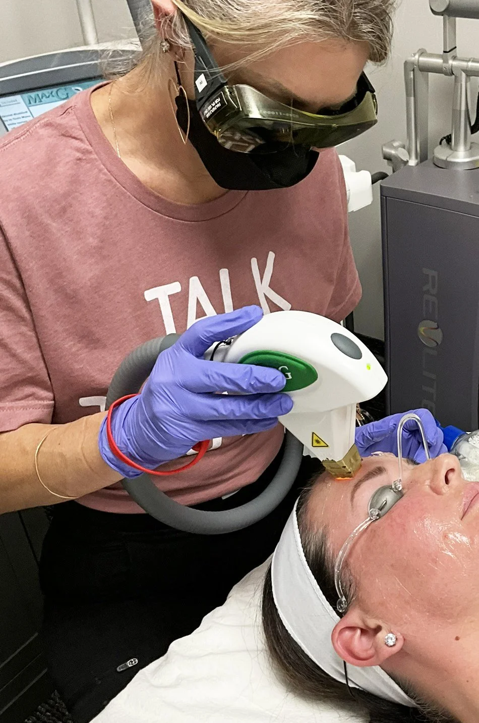 A person wearing protective goggles and gloves performs a laser skin treatment on a woman's face, who is lying down with a bandage around her head and wearing an oxygen tube.