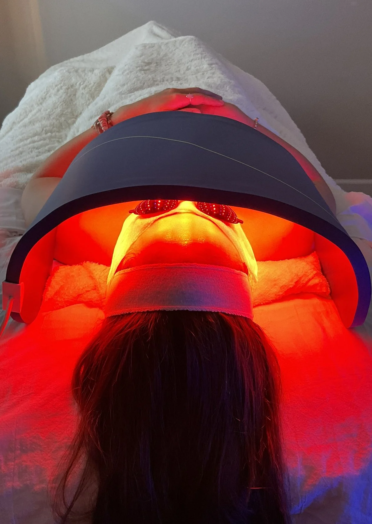 Person lying on a table under a red LED light therapy device, wearing protective glasses, with long dark hair and a yellow towel around their neck.