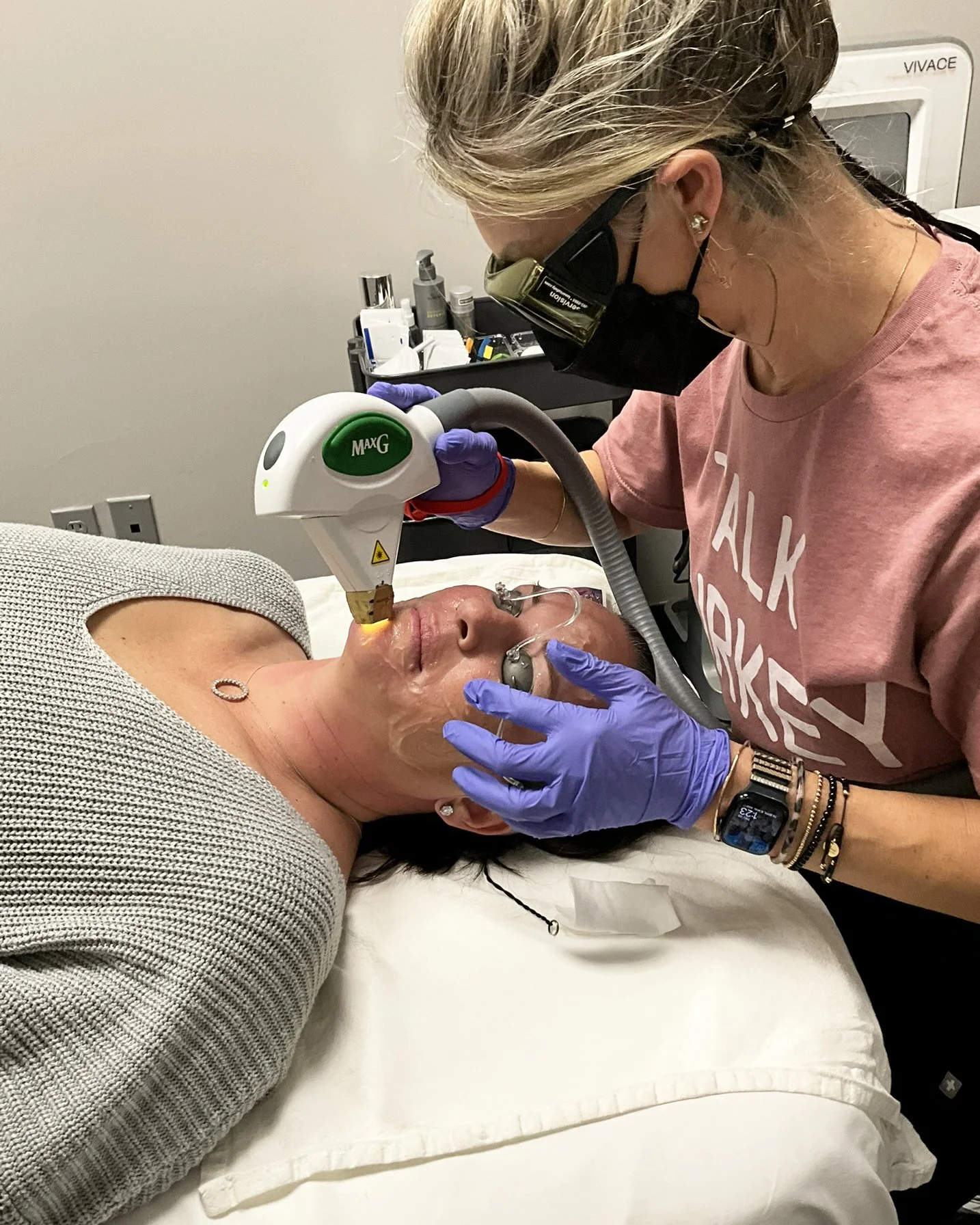 A woman receiving a facial treatment with a laser device in a medical setting, wearing purple gloves, a pink T-shirt, a black face mask, and protective eyewear.
