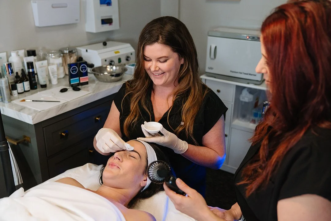 A woman lying on a treatment bed with a headband, receiving a facial treatment from two women in a spa or clinic setting. One woman is using a handheld device while the other holds a skincare tool, and they are smiling.