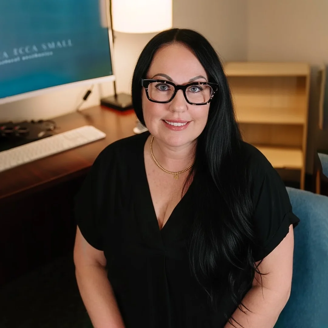 A woman with long black hair, wearing glasses and a black top, sitting in a room with a computer, lamp, and wooden shelving behind her.