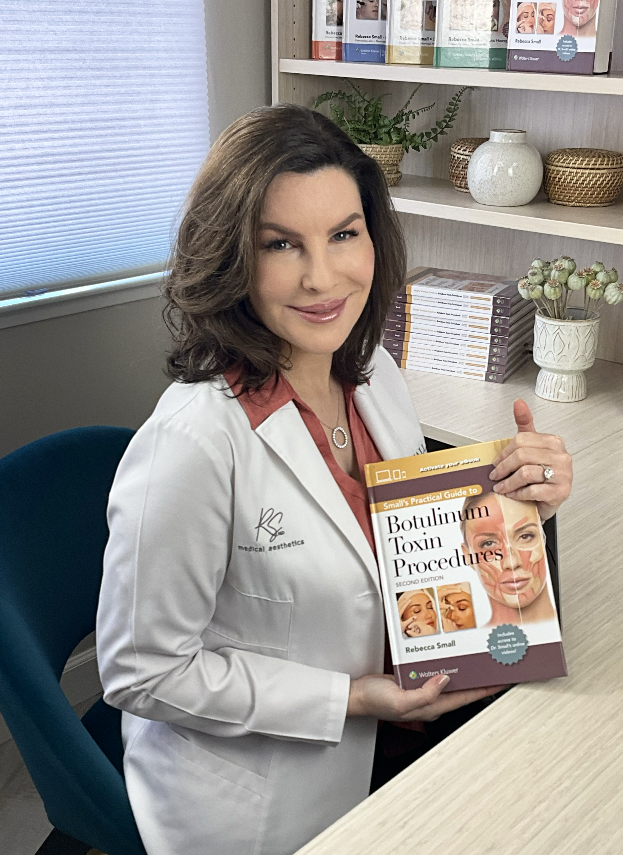 A woman in a white medical coat sitting at a desk, holding a book titled 'Botulinum Toxin Procedures' by Rebecca Small. She is smiling and seated in an office with books and decorative items on shelves behind her.