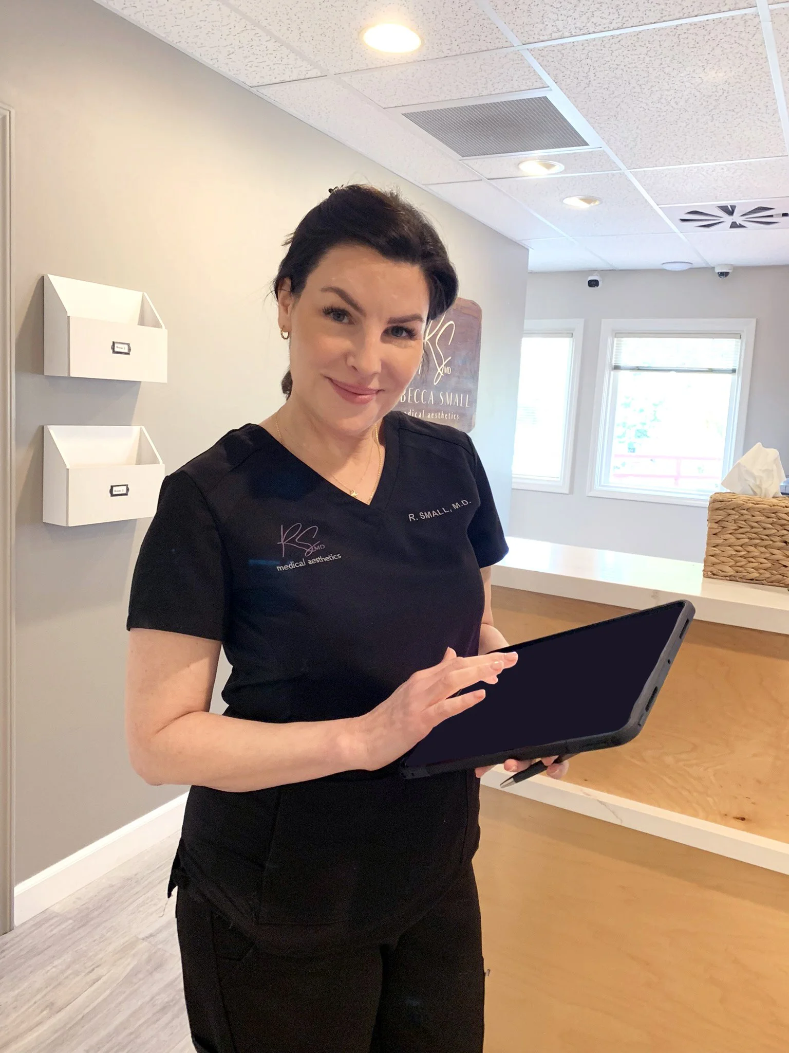 A woman wearing a black medical uniform standing indoors, holding a tablet. There are windows and a counter in the background.