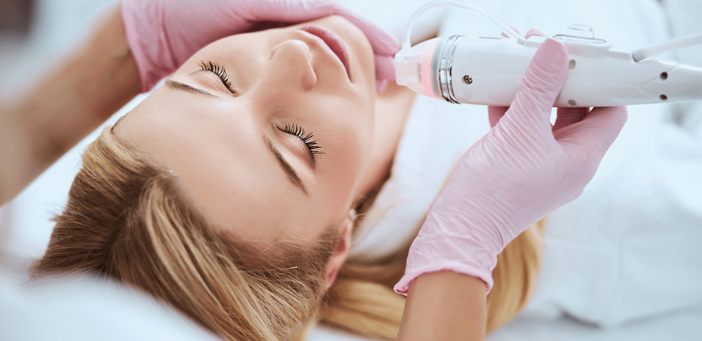 A woman lying down during a cosmetic facial treatment with a dermal vacuum device, wearing pink gloves.