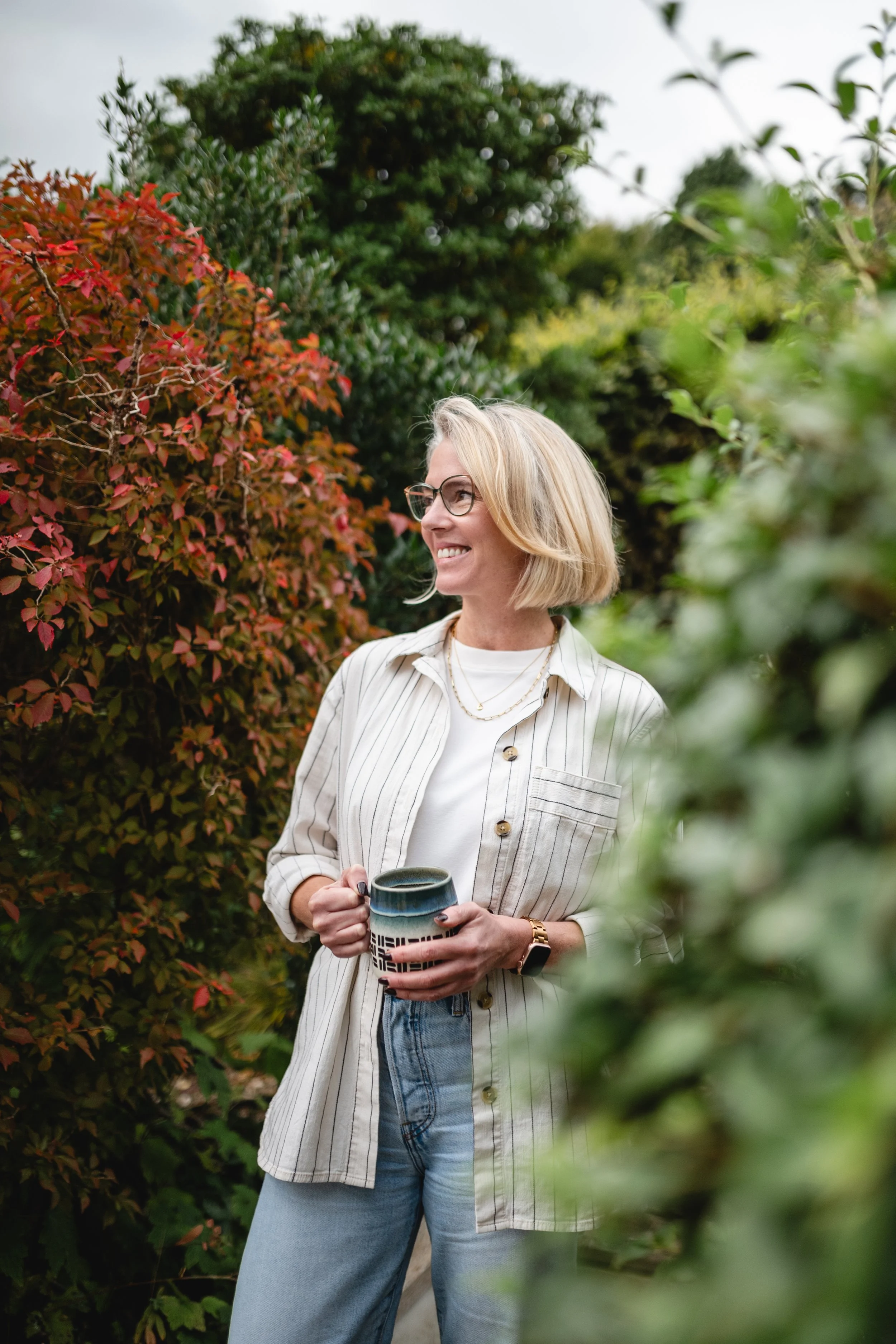 A woman with blonde hair and glasses smiling while holding a mug, standing outdoors among greenery and red foliage.