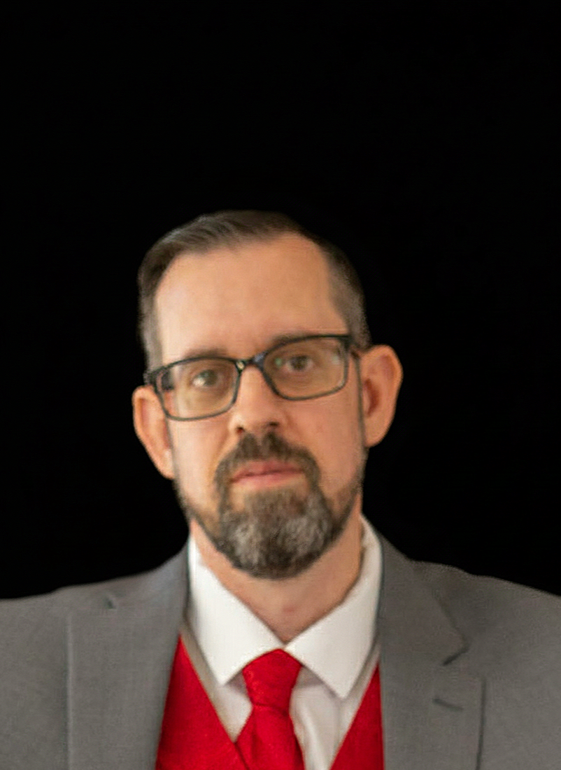 Portrait of Andrew Hersey, co-owner of Hersey Creative Studios, wearing a gray suit and red tie against a black background