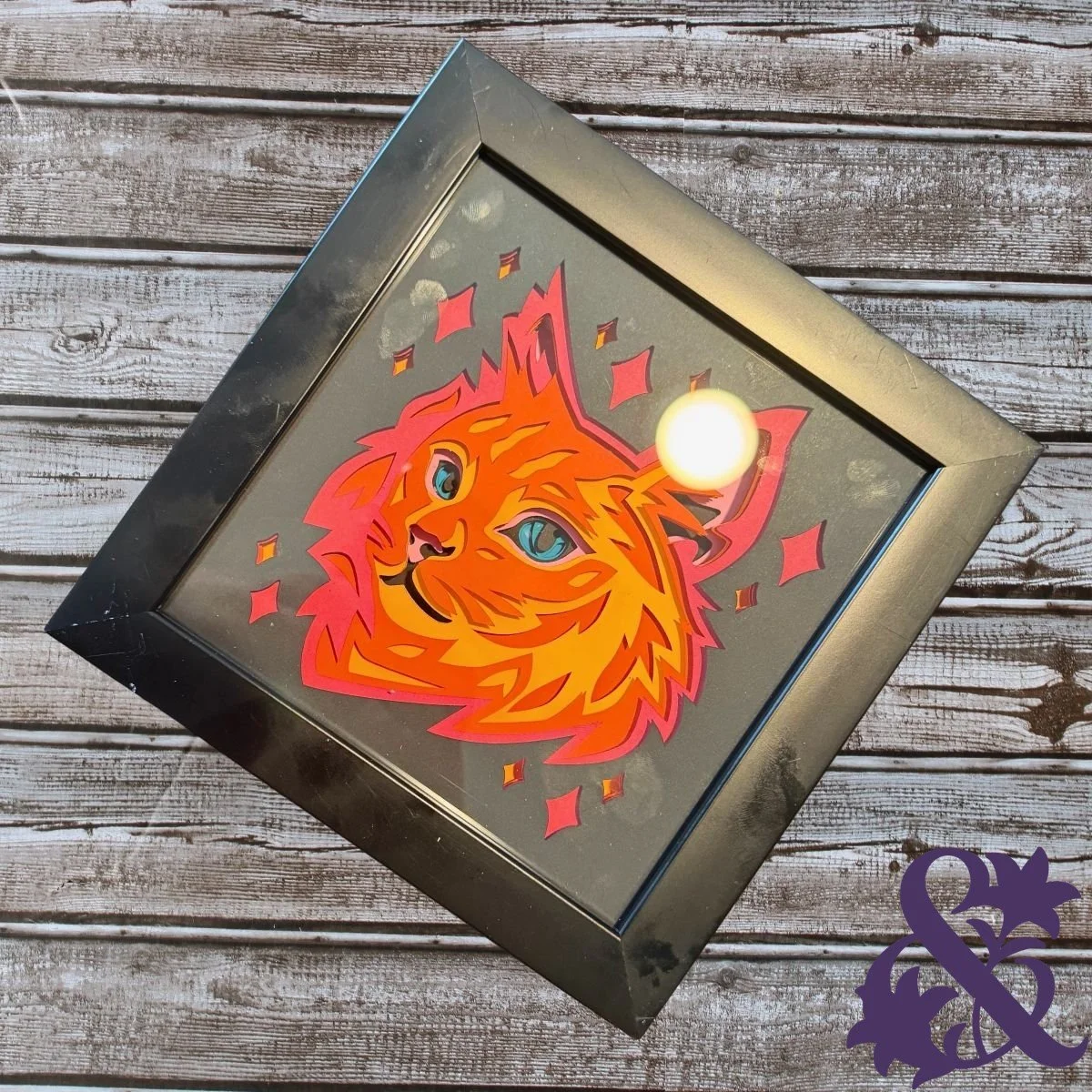 Framed layered-paper artwork featuring a bright orange cat with blue eyes