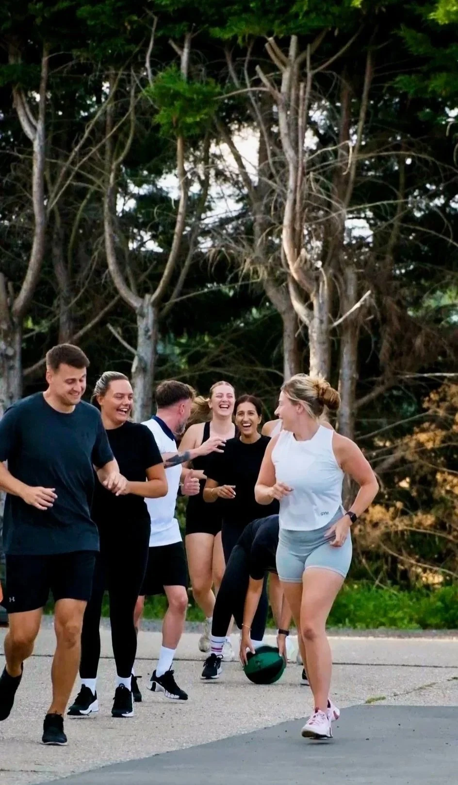 Group of people jogging and laughing outdoors in a park, with trees in the background.