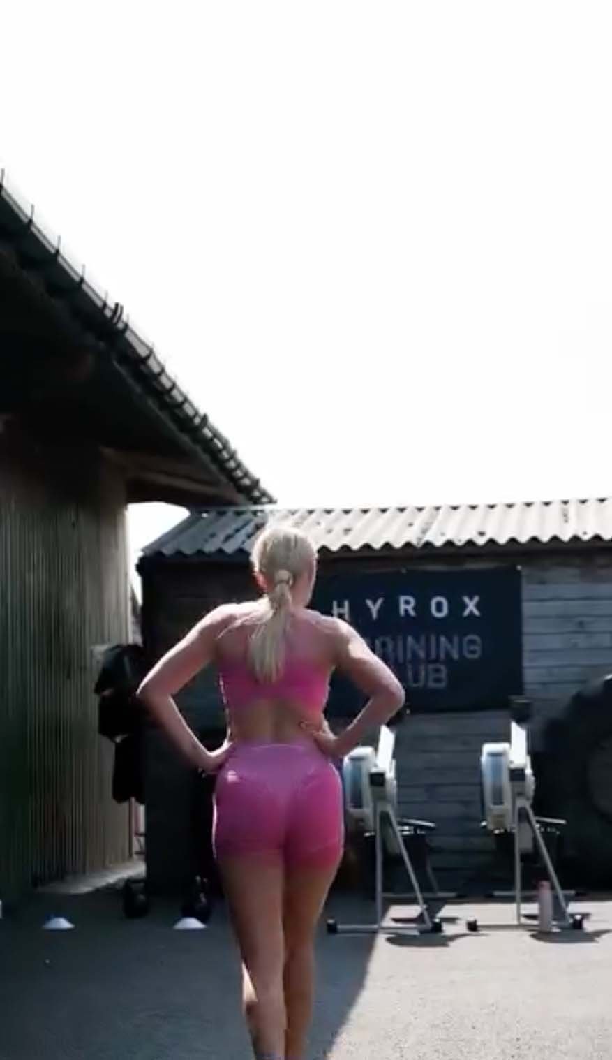 Woman with blonde hair tied back in a ponytail, wearing a pink sports bra and pink shorts, walking away from the camera at an outdoor gym area with fitness equipment and a sign that reads 'HYROX TRAINING CLUB'.