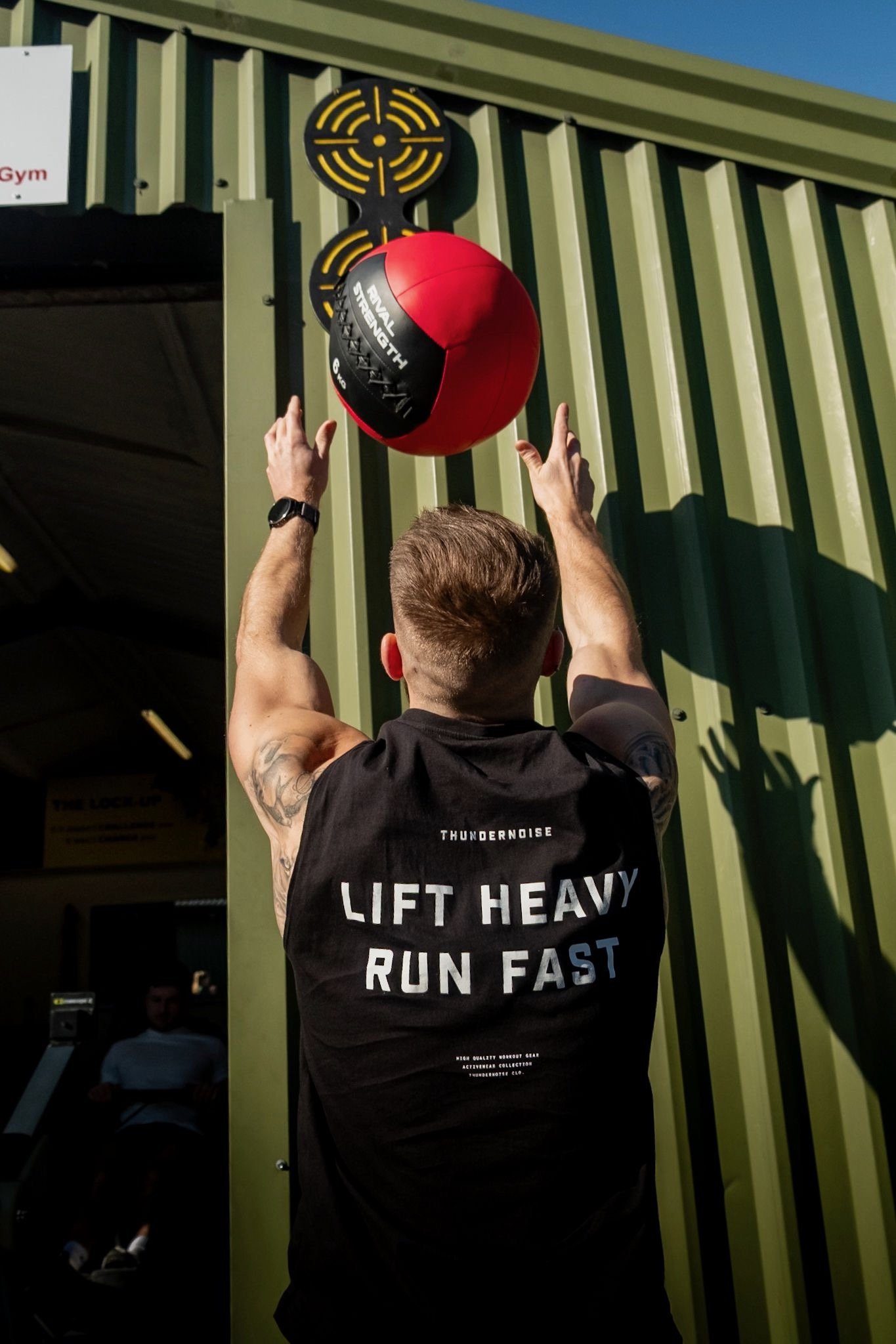 A man in a black tank top with the words 'Lift Heavy Run Fast' on the back is throwing a red medicine ball against a green wall at a gym. The ball is hitting a target above.