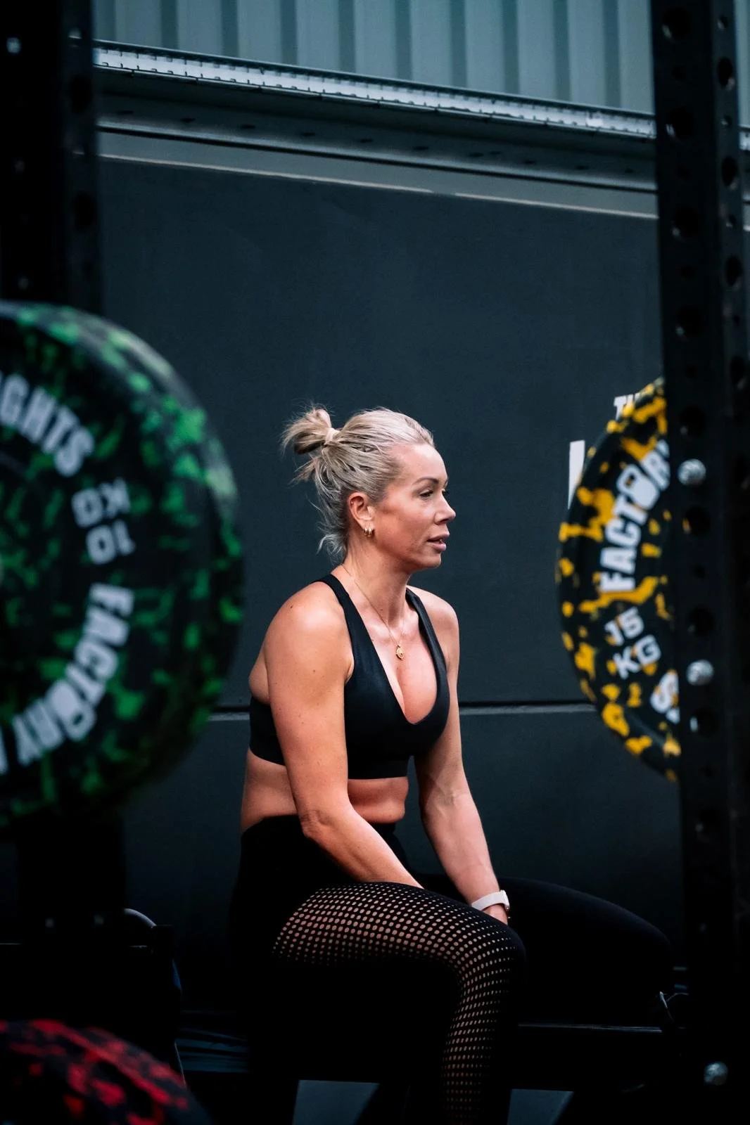 A woman in workout attire sitting in a gym, surrounded by weight plates, with her hair tied back and a focused expression.
