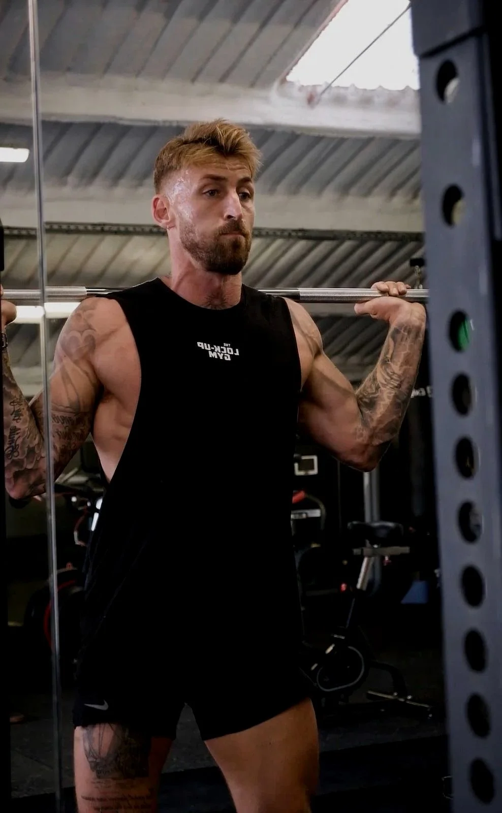 A man with tattoos and a beard lifts a barbell during a workout at a gym.