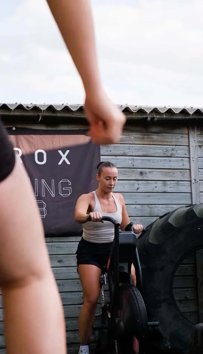 Woman working out on an indoor rowing machine in a gym with a large tire to her right and a wooden wall behind her.