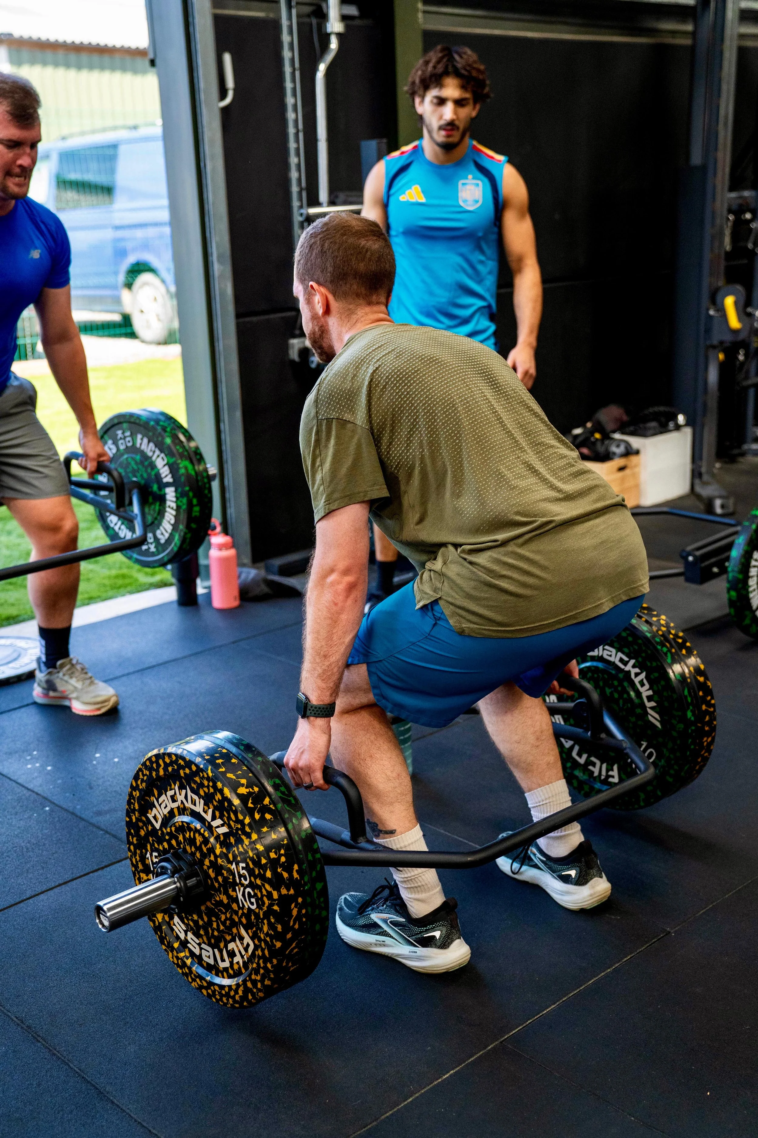 A man in a green shirt and blue shorts performing a deadlift with a barbell loaded with black and yellow weight plates, in an indoor gym. Two other men are present, one in a blue shirt and gray shorts, and the other in a blue athletic tank top.