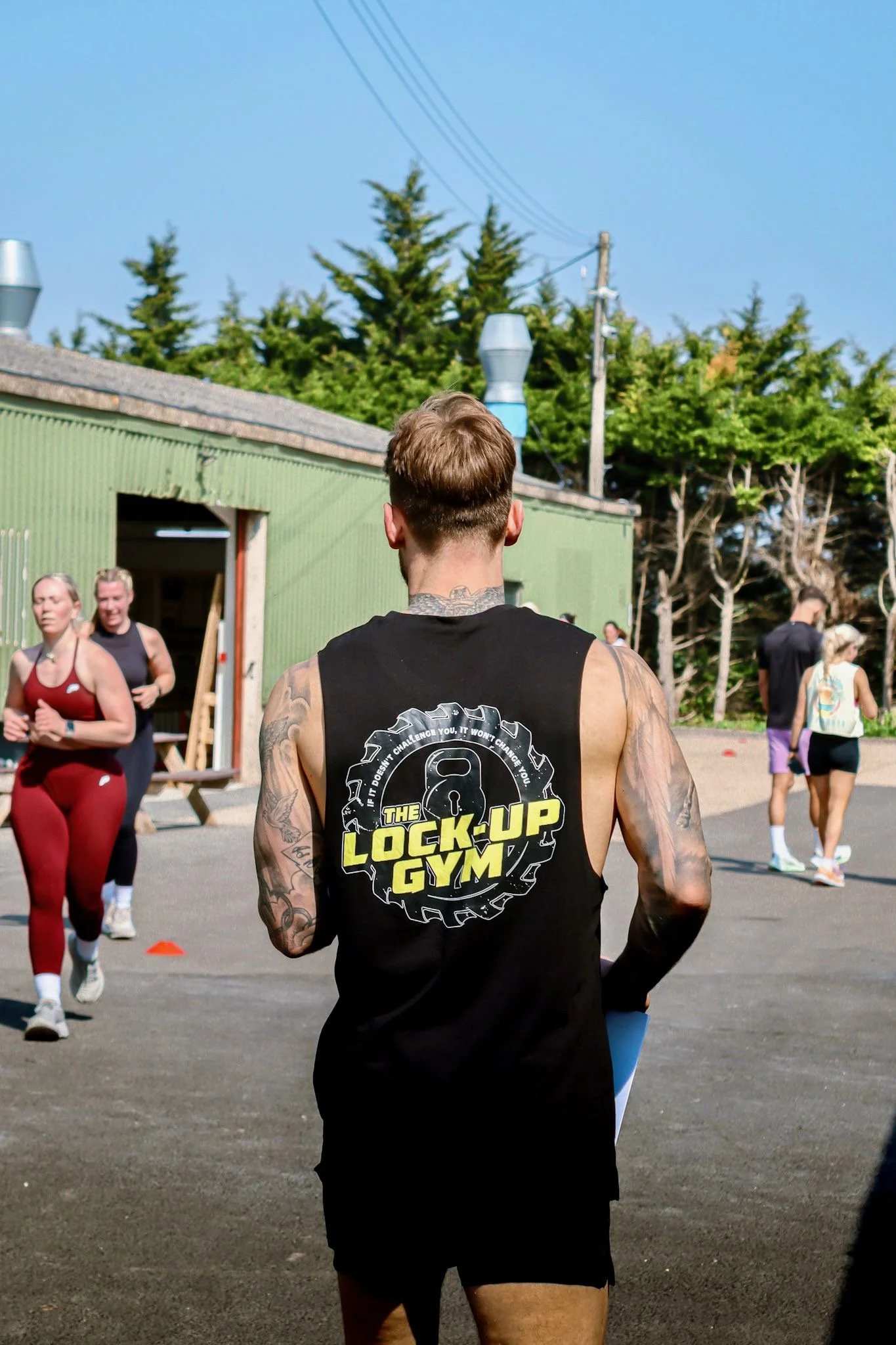 Back view of a man with tattoos wearing a black tank top with yellow and white text that reads 'The Lock-Up Gym', standing outdoors with people running and walking in the background under a clear blue sky.