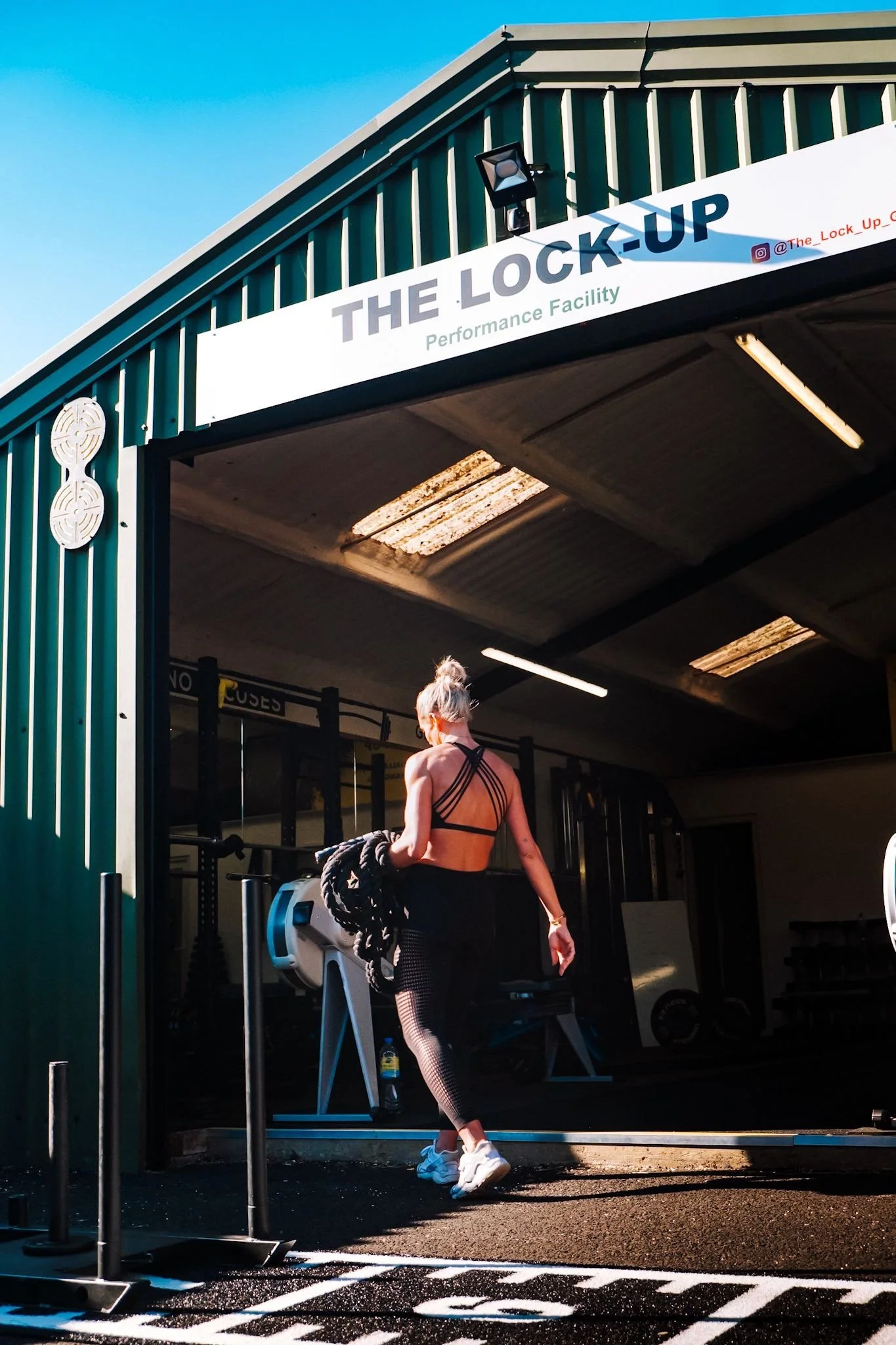 Woman with blonde hair tied up, wearing black workout clothes, carrying battle ropes, walking into a fitness gym named The Lock-Up