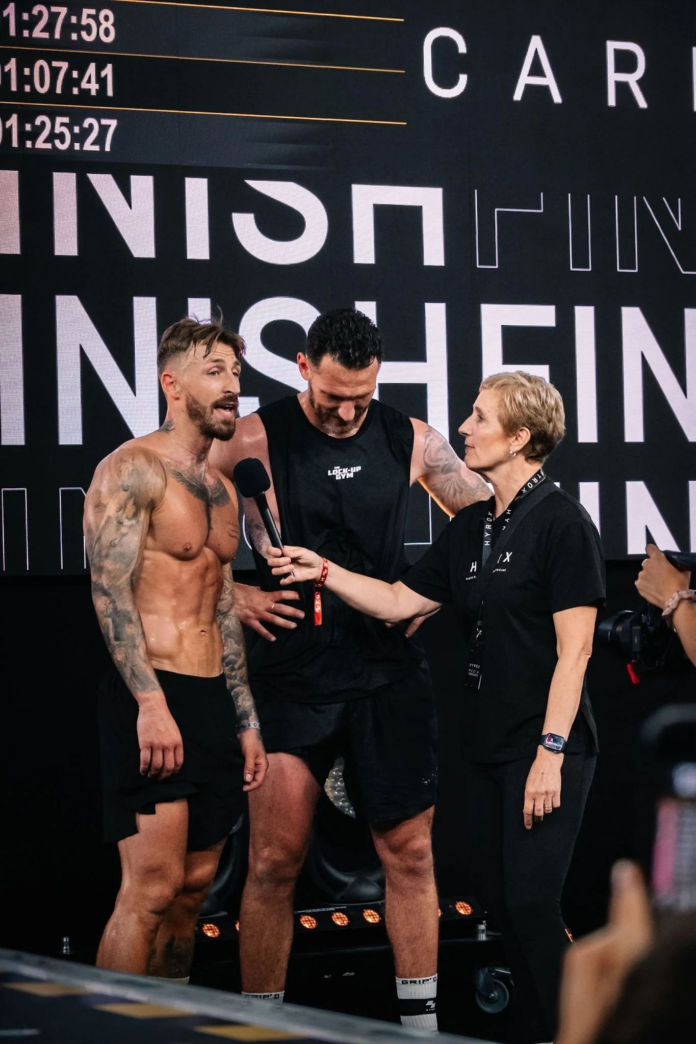 Three people on stage, one shirtless man with tattoos being interviewed with a microphone, another man in black athletic wear, and a woman with short blonde hair holding the microphone. A digital display in the background shows race times.
