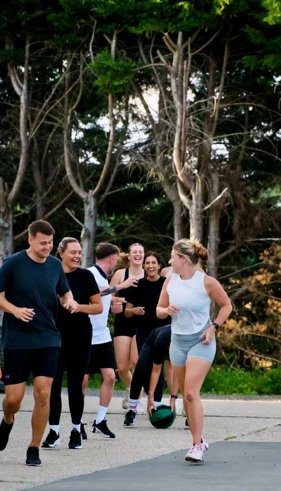 Group of people in athletic clothing smiling and laughing while exercising outdoors near trees, with some using a medicine ball.