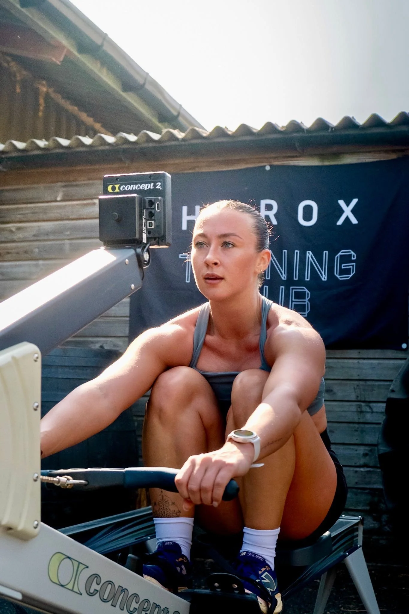 A woman exercising on a rowing machine outdoors, with a black banner behind her that reads 'HEROX TRAINING CLUB'. She is wearing a gray sports bra, black shorts, and a white watch, with her hair tied back.
