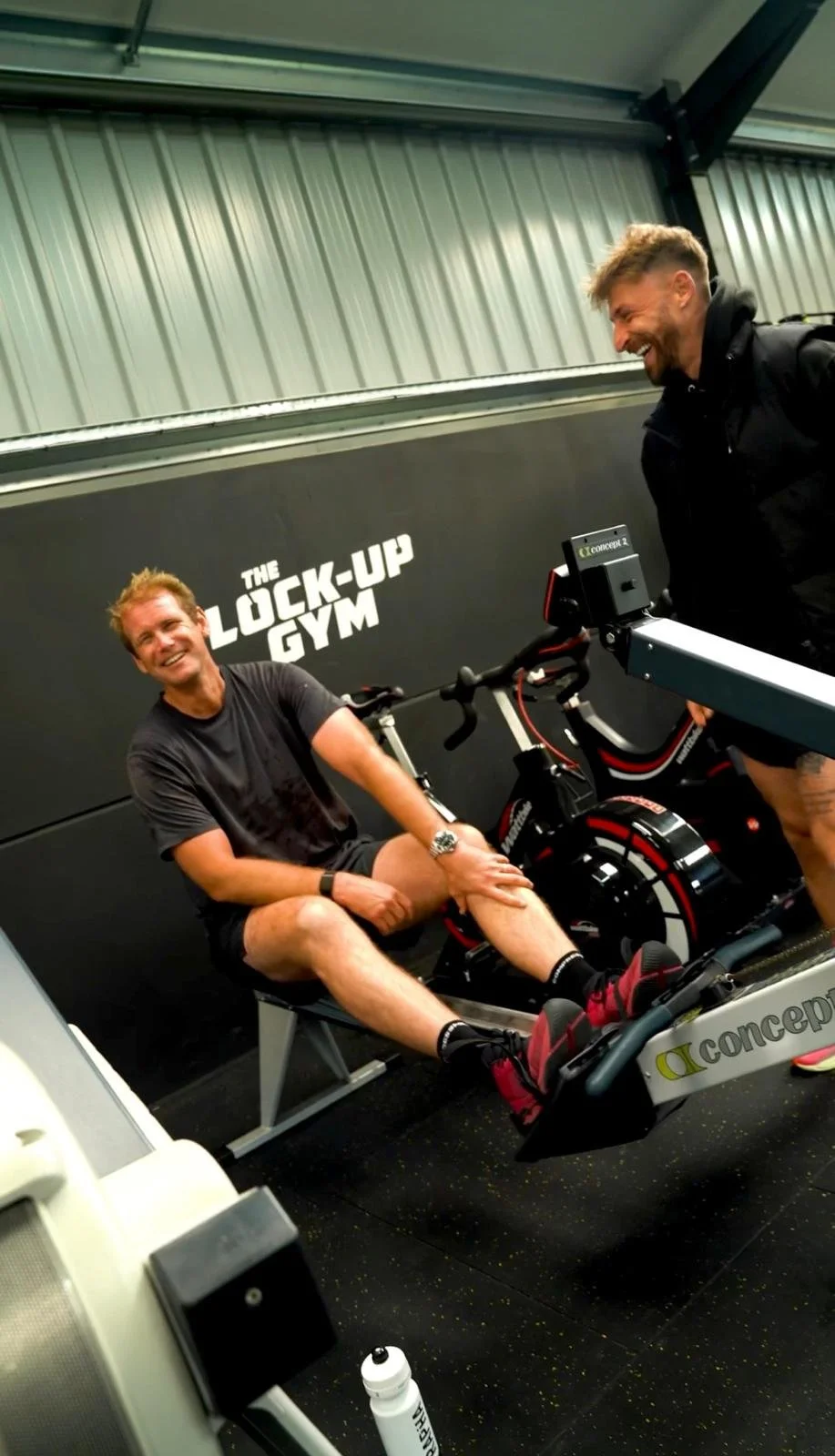 Two men at The Lock-Up Gym with one on a stationary exercise bike, smiling and talking.