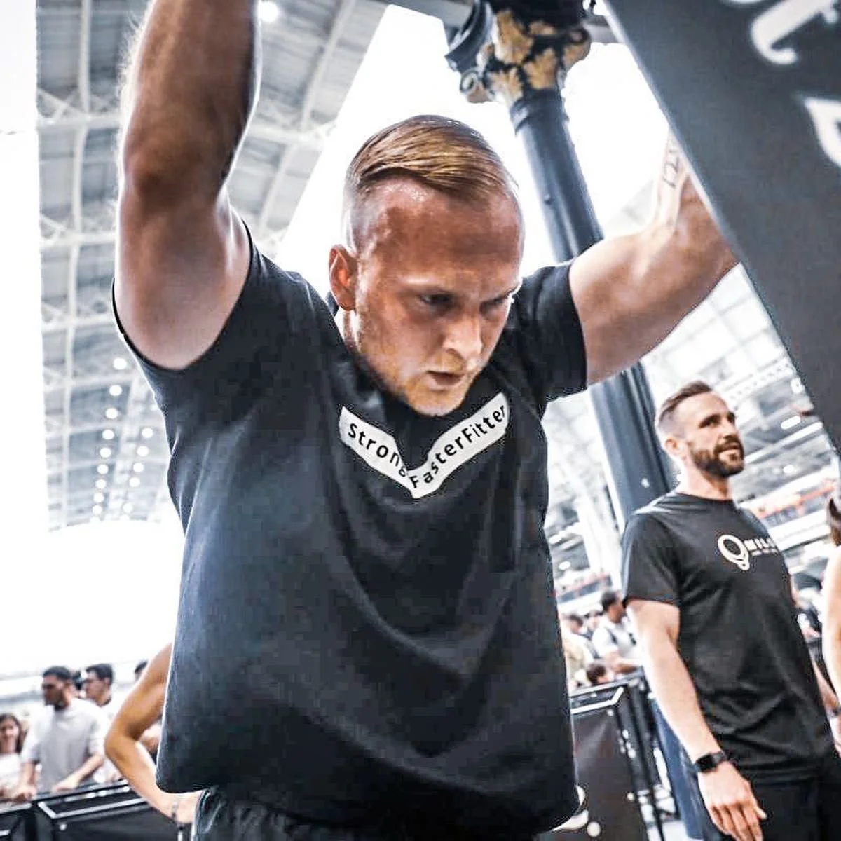 A man with short hair and a beard wearing a black shirt with the words 'Stronger FasterFitter' engaged in a workout at a gym, with another man in a similar black shirt observing in the background.