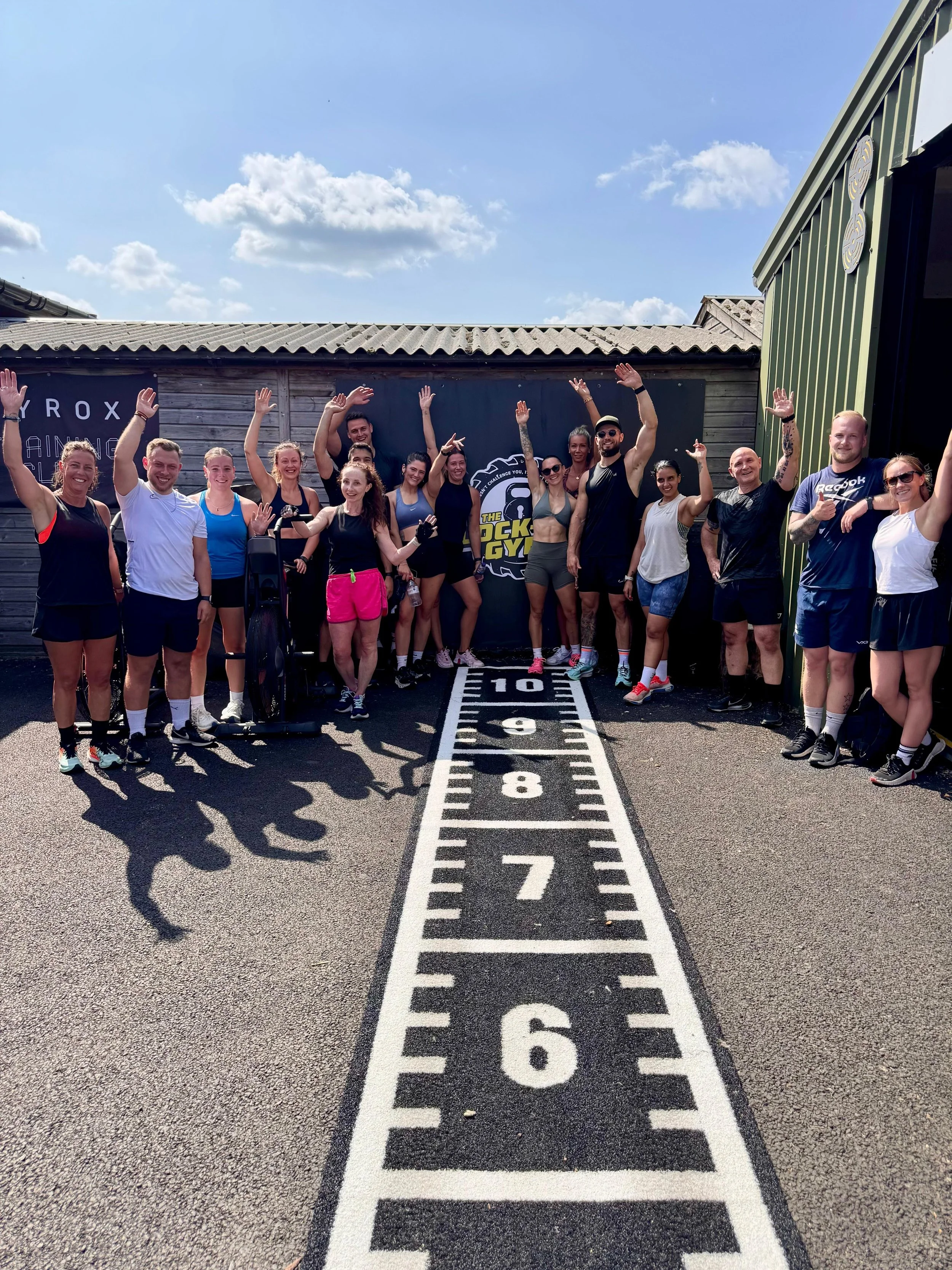 Group of people standing outdoors on a fitness track with their arms raised and smiling. They are dressed in workout clothes, with some holding water bottles, under a blue sky with some clouds. There are signs and a green building in the background.