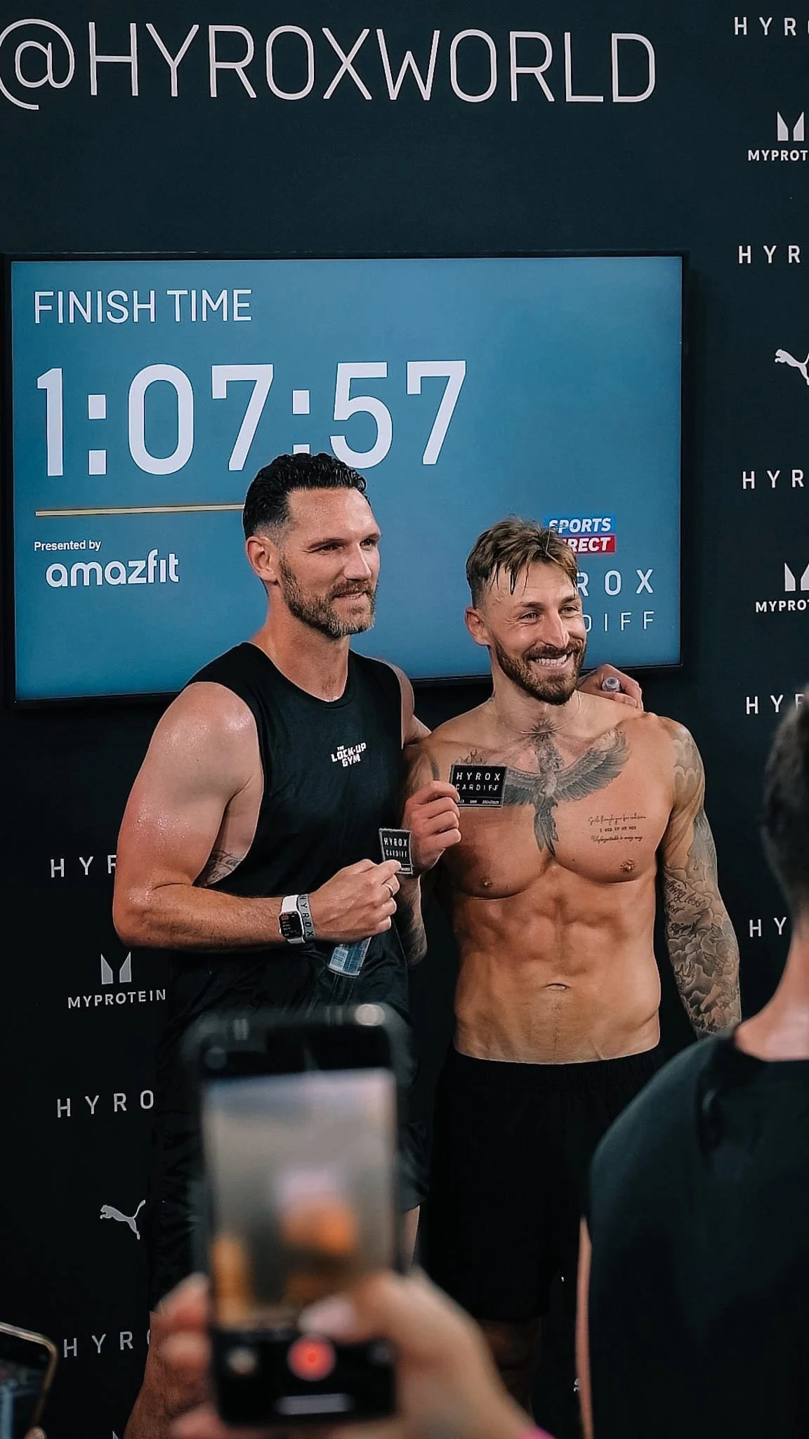 Two men celebrating after a race, one shirtless with tattoos, the other in a black sleeveless shirt, holding a medal with a timer showing 1:07:57 behind them.