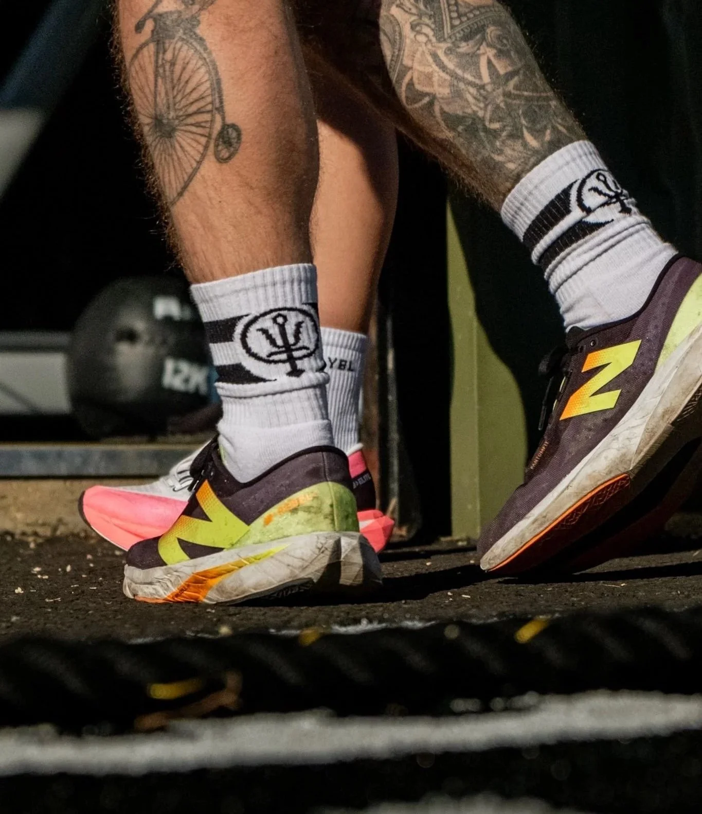 Close-up of a person's legs in white athletic socks with a logo, wearing black and yellow New Balance running shoes, positioned on a black textured floor, with a fitness equipment in the background.