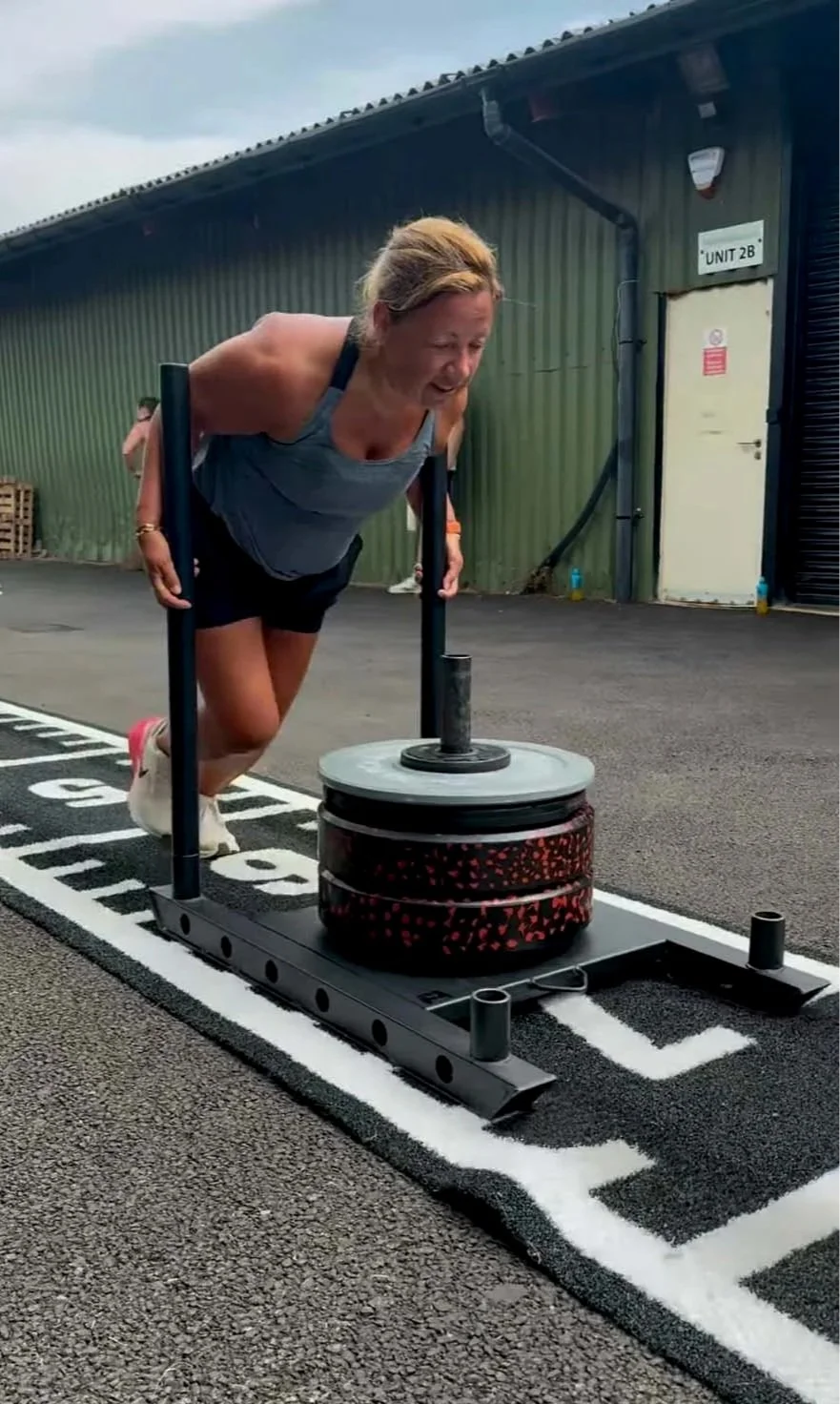 Woman in athletic wear doing a push-up on a weighted sled outside, with a green industrial building in the background.