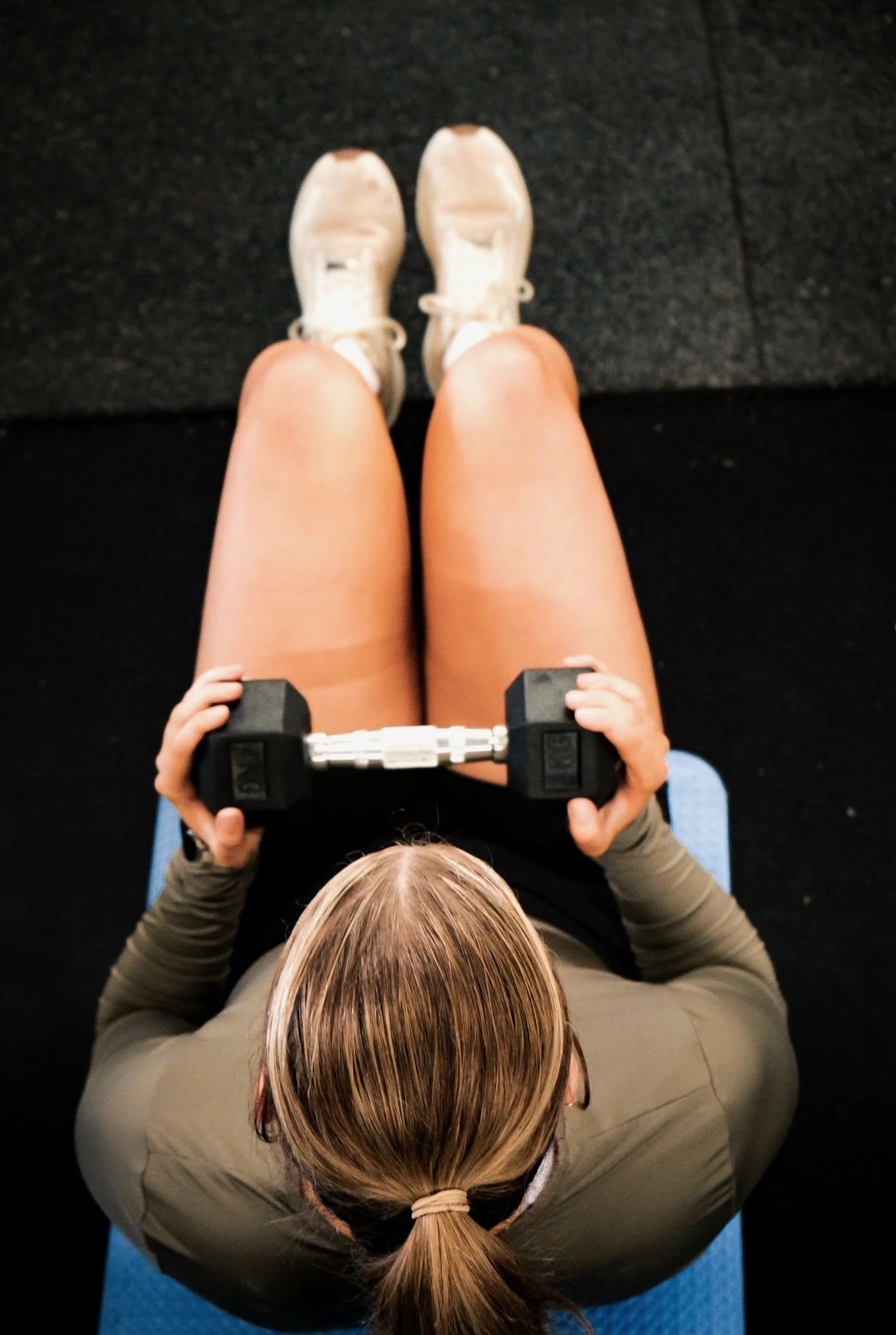 A person sitting on a blue exercise mat, holding a small dumbbell with both hands, wearing white sneakers, tan shorts, and a gray long-sleeve shirt, with hair tied back in a ponytail.