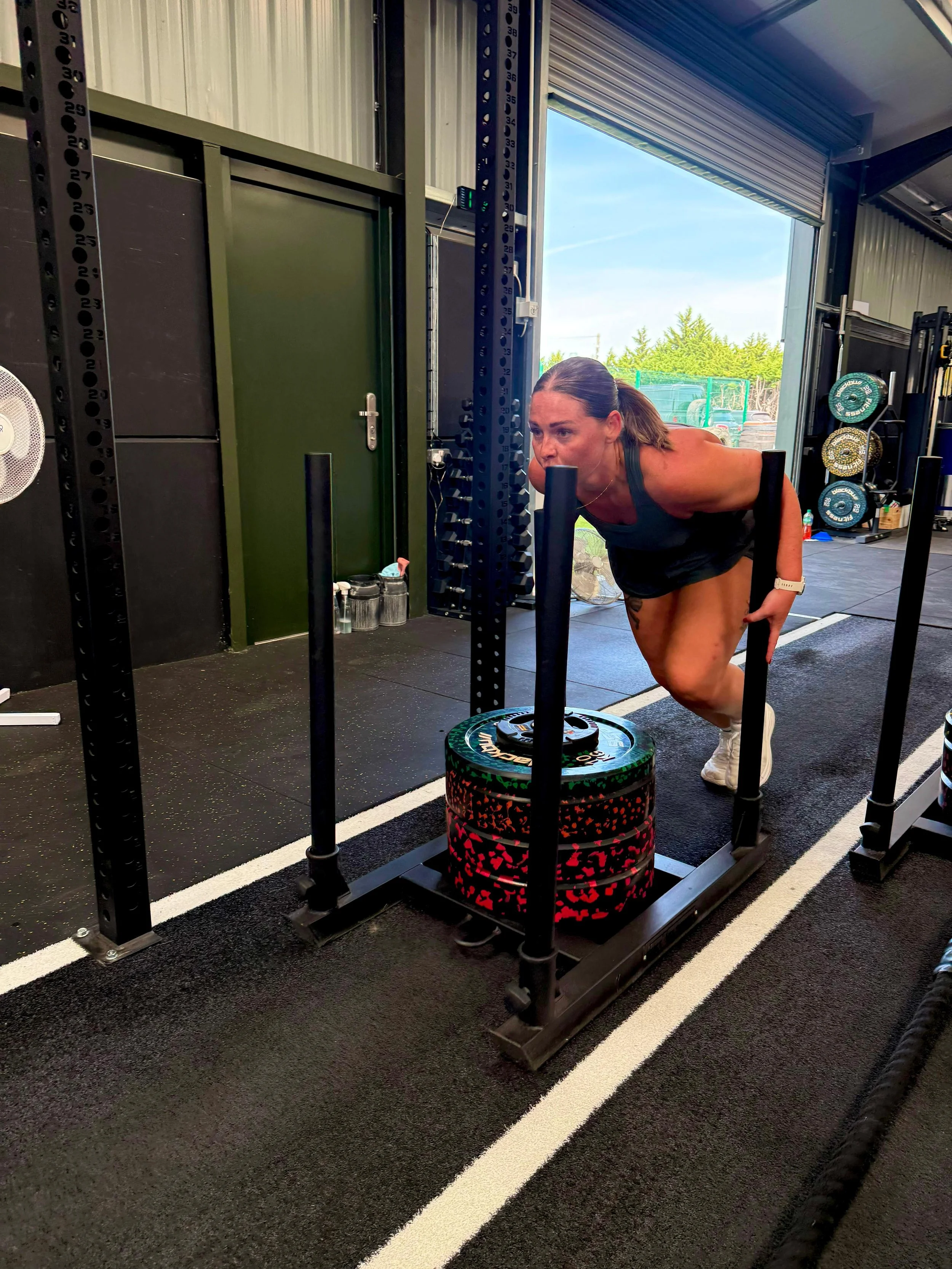 A woman performing a tire flip exercise in a gym, with a black equipment rack, weights, and an open garage-style door showing trees outside.