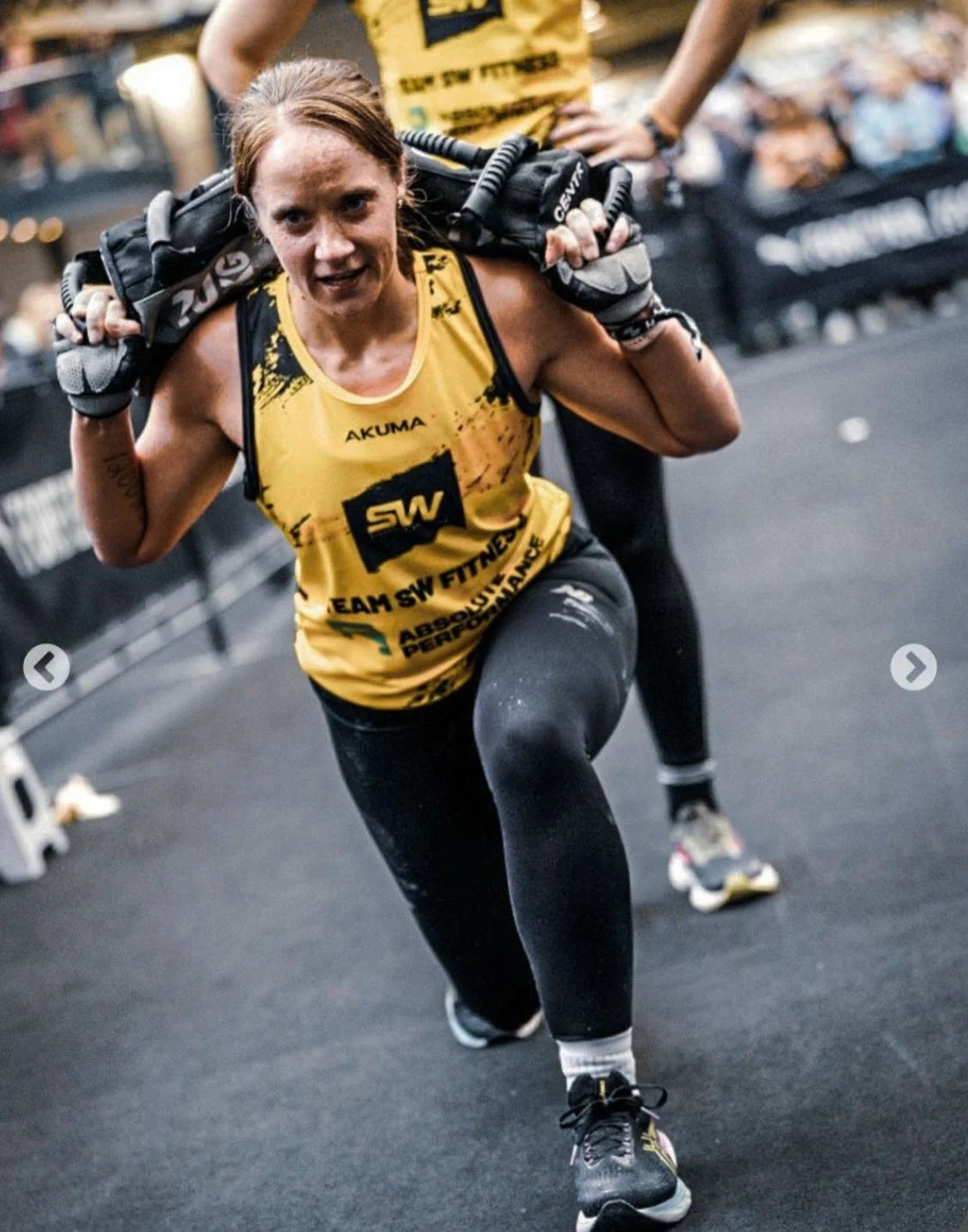 Female athlete in yellow tank top and black leggings doing a weighted lunge exercise with a black sandbag on her shoulders in a gym.