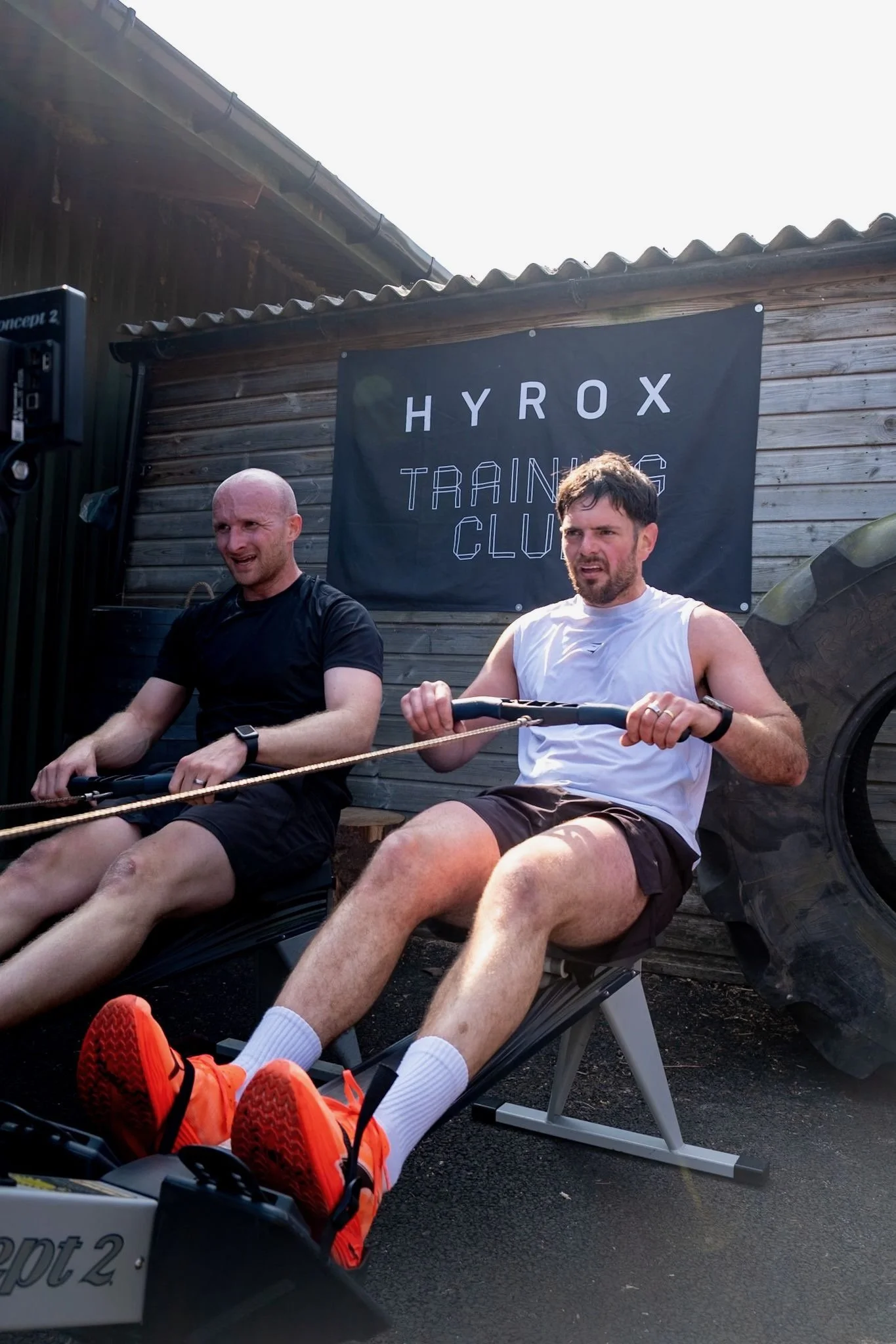 Two men exercising on rowing machines outdoors in front of a wooden wall with a Hyrox Training Club banner, a large tire, and a clear sky.