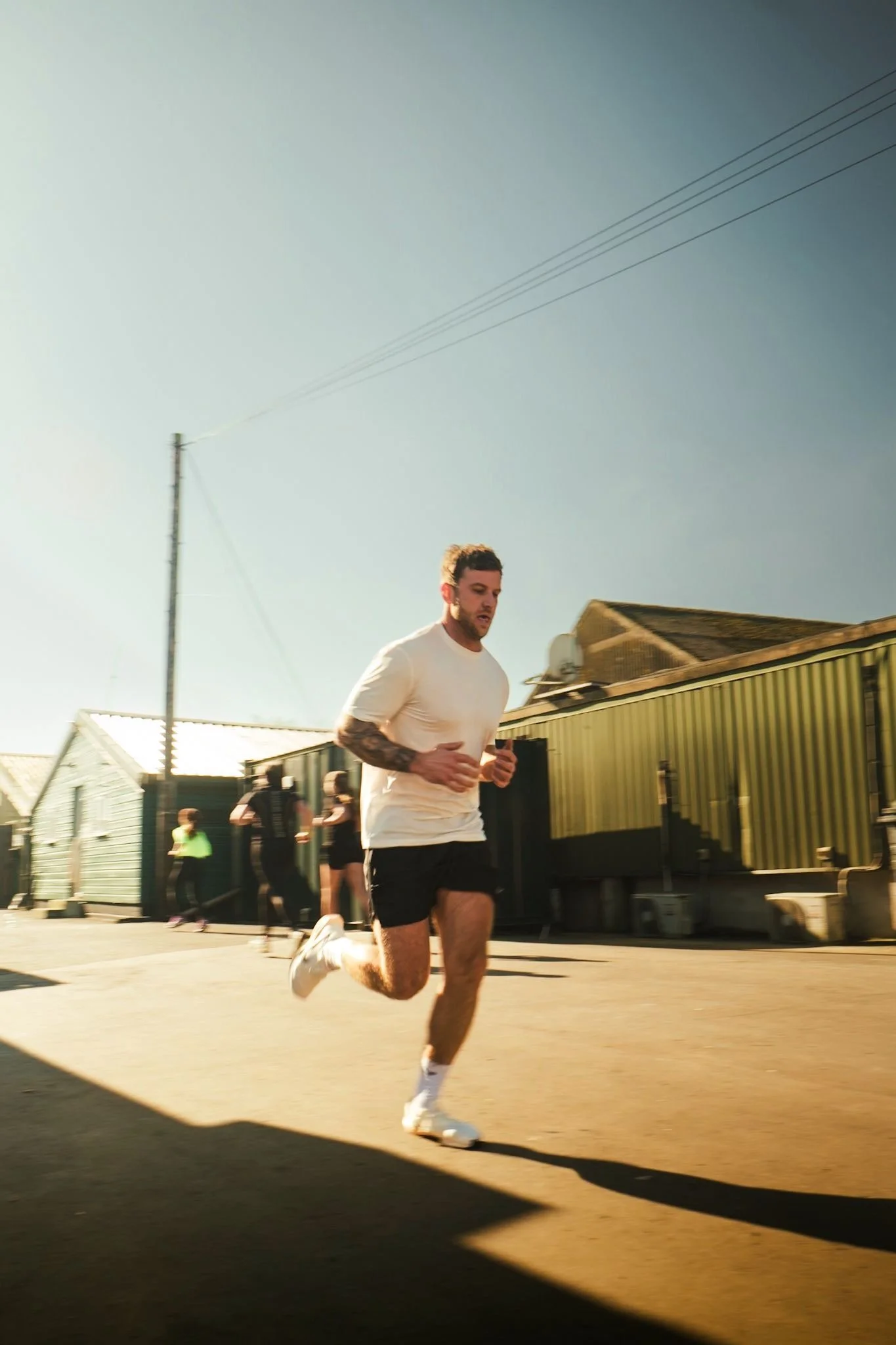 A man running outdoors on a sunny day, wearing a white t-shirt, black shorts, and white running shoes, with a tattooed arm and several people in the background near green and yellow buildings.