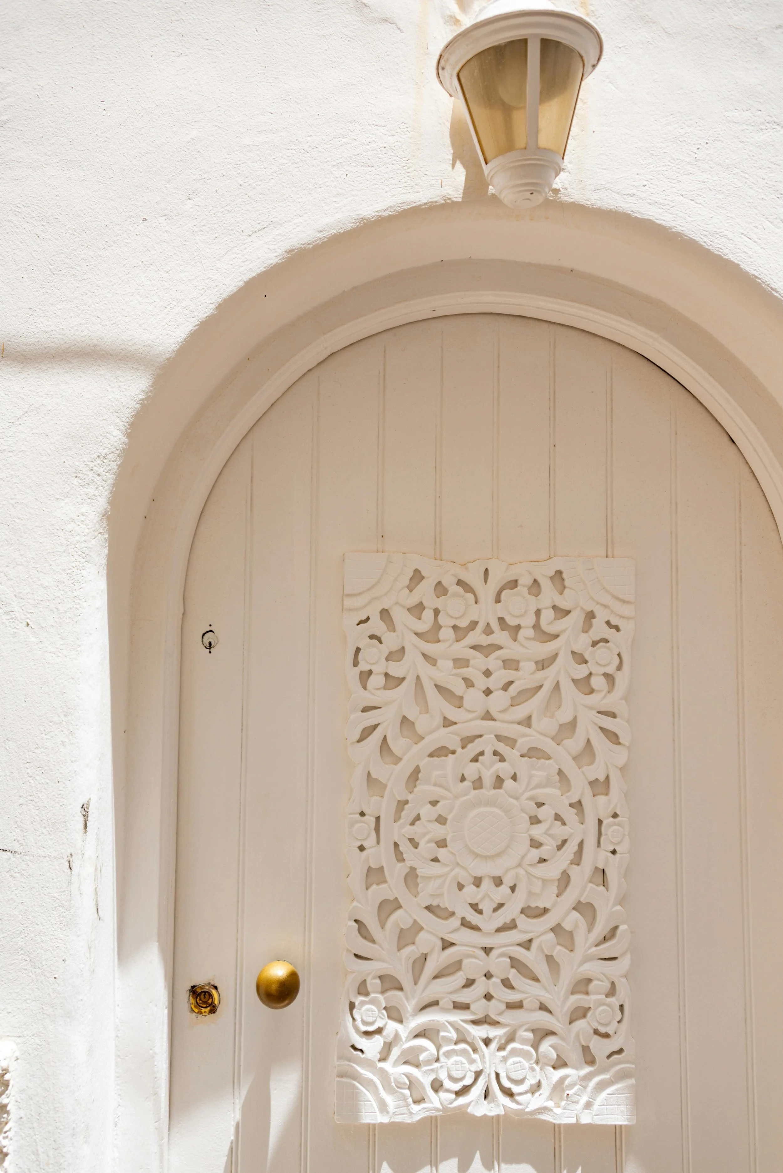 A white front door with ornate carved decoration, a golden doorknob, a small hole where a lock might go, and a light fixture above the door. The door is set within a white textured wall.