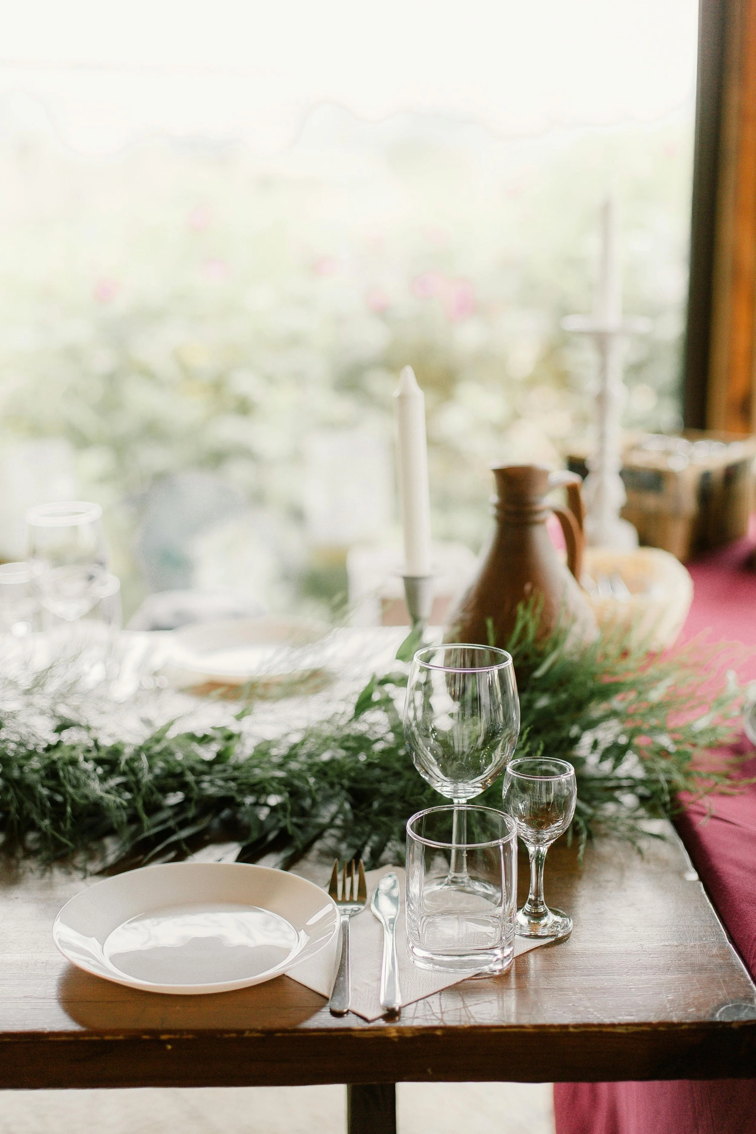 Elegant table setting with a white plate, silverware, clear glassware, and a green leafy centerpiece on a wooden table, with a blurred window and outdoor scenery in the background.