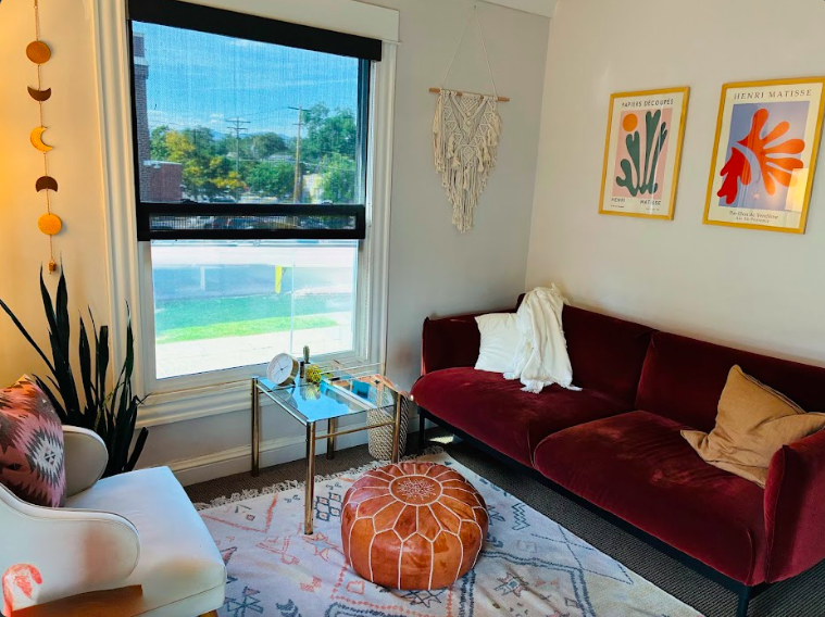 Living room with a large window, a white armchair with a patterned pillow, a burgundy velvet sofa with pillows, a round leather pouf, a glass-top side table with decor, and wall art featuring botanical prints.