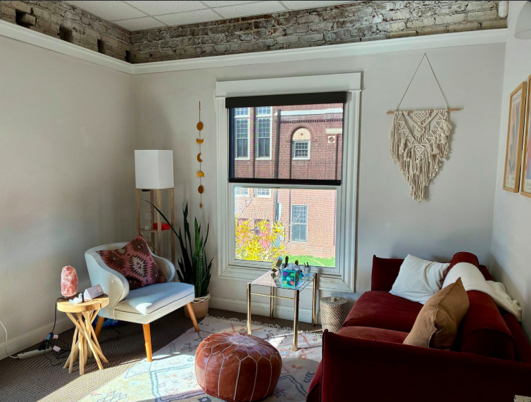 Cozy living room with white armchair, red sofa, wooden side table, and large window showing brick buildings outside. Decor includes hanging wall art, floor lamp, houseplants, and a pouf in front of the sofa.