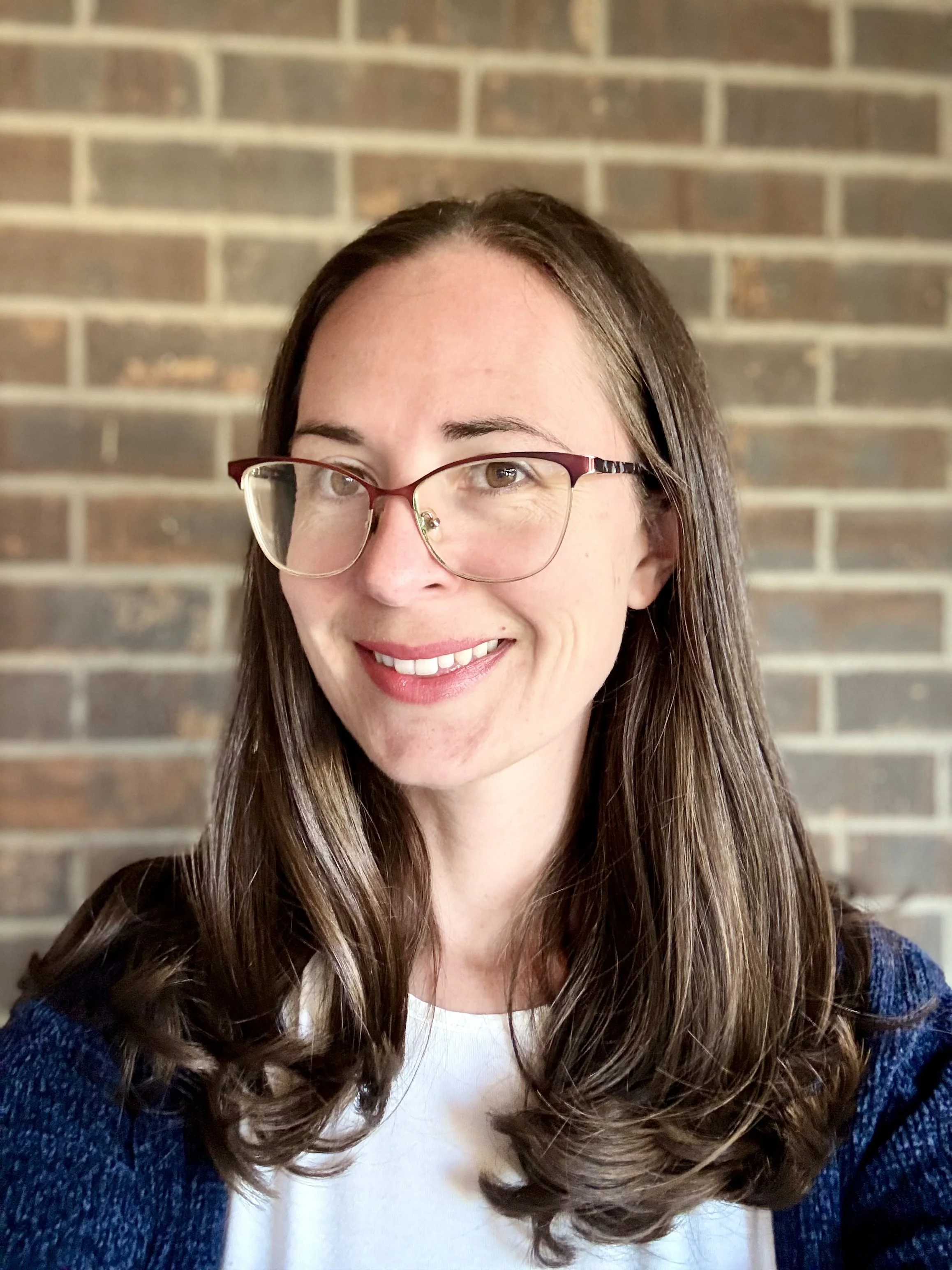 A woman with glasses and long brown hair smiling in front of a brick wall.