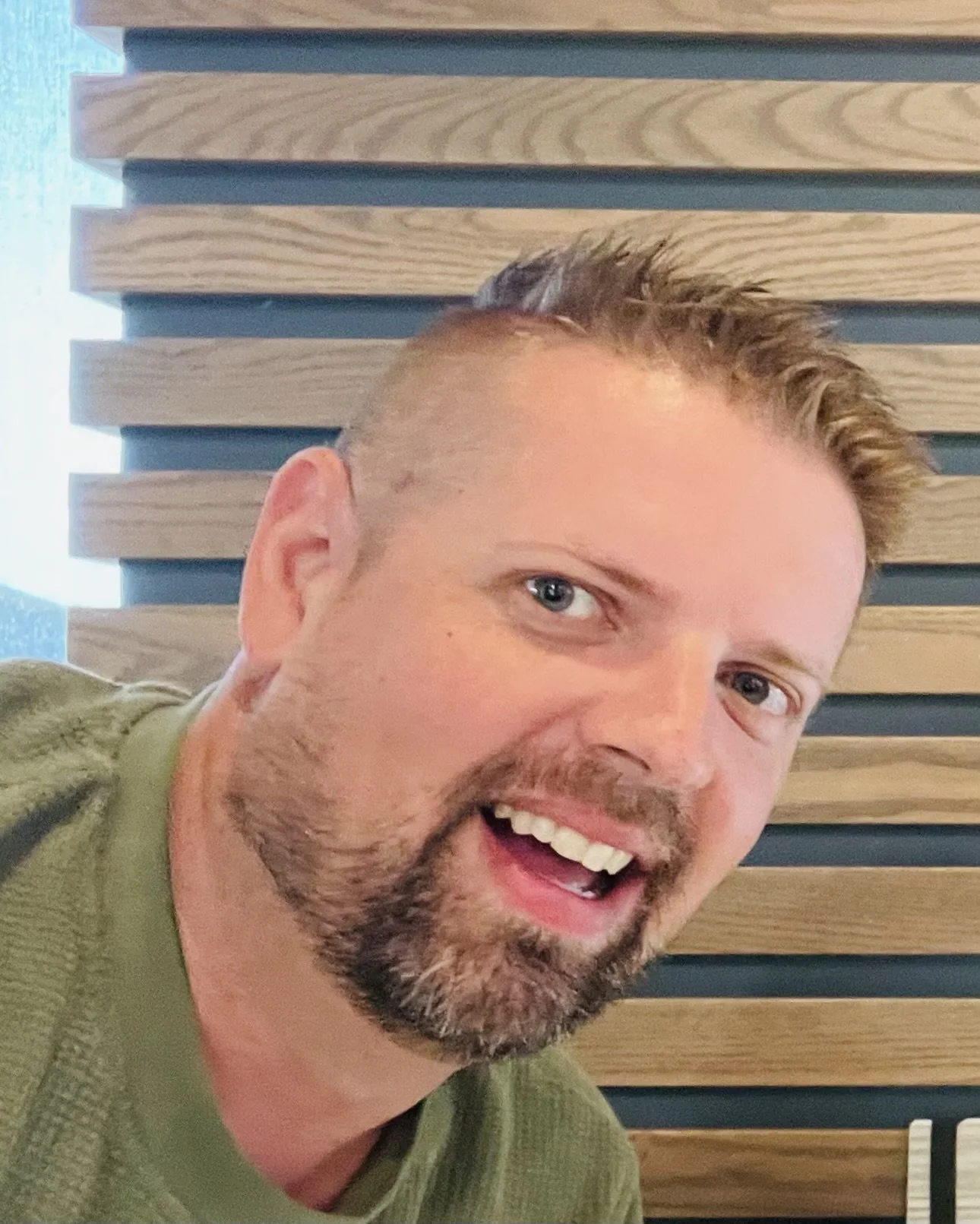 A man with short, light brown hair, a beard, and blue eyes, smiling with his mouth open, wearing a green shirt, with a wooden slatted wall in the background.