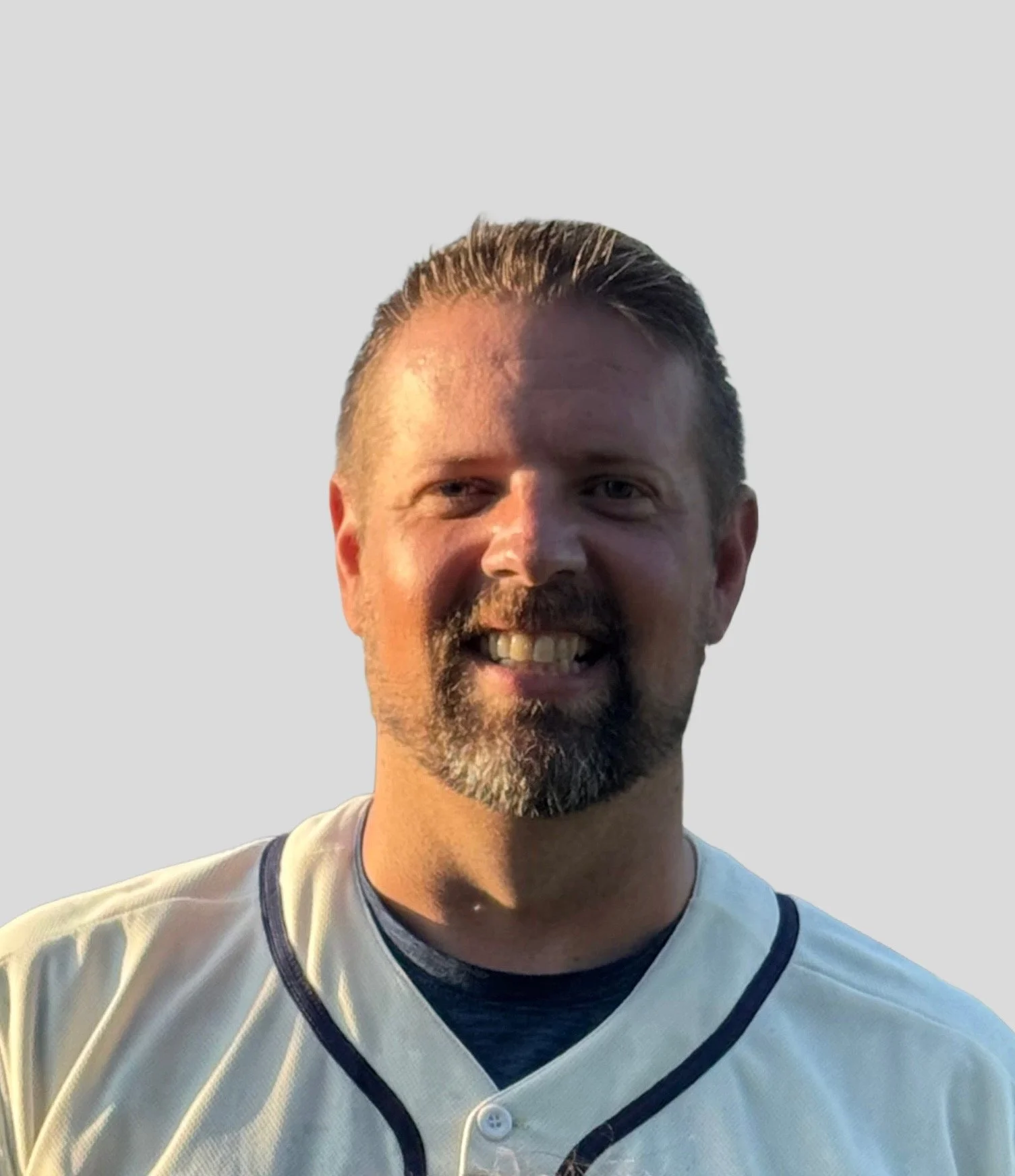 A smiling man with a beard and mustache, wearing a white sports jersey with black trim, standing against a light background.