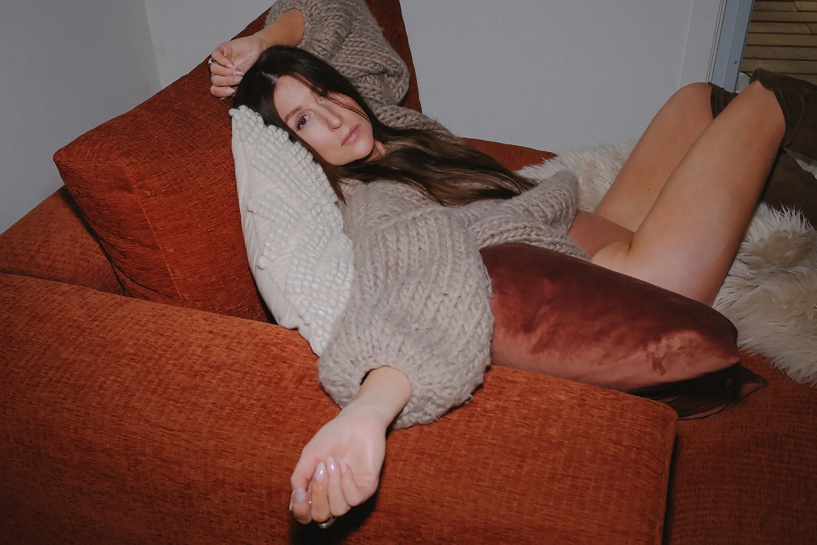 Woman lying on a reddish-brown couch, wearing a chunky beige sweater, with a white pillow under her head, and reaching with her left hand while looking into the camera.