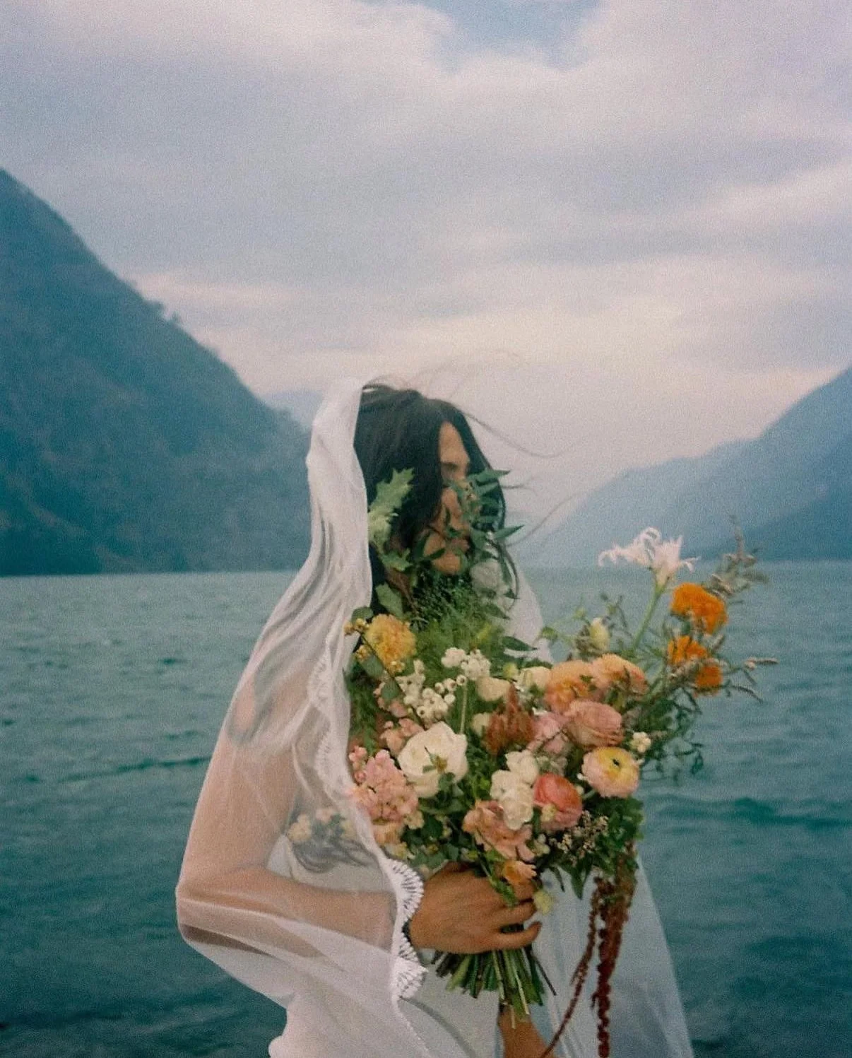 A woman dressed in a white veil and lace robe holding a bouquet of colorful flowers by a body of water with mountains in the background.