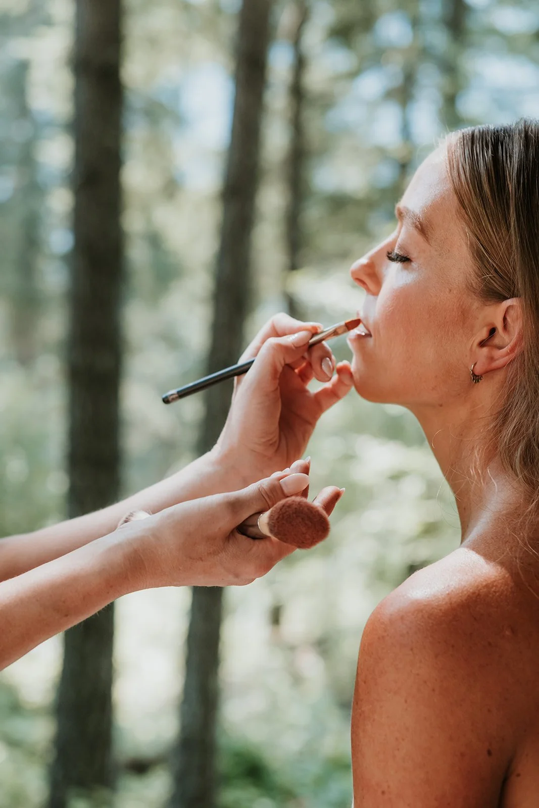 A woman with fair skin and freckles is having makeup applied in a forest setting with tall trees in the background.